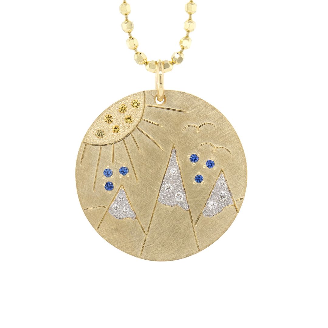 14k Gold round pendant with mountain etching with white diamond snow capped mountains and yellow and blue sapphires.