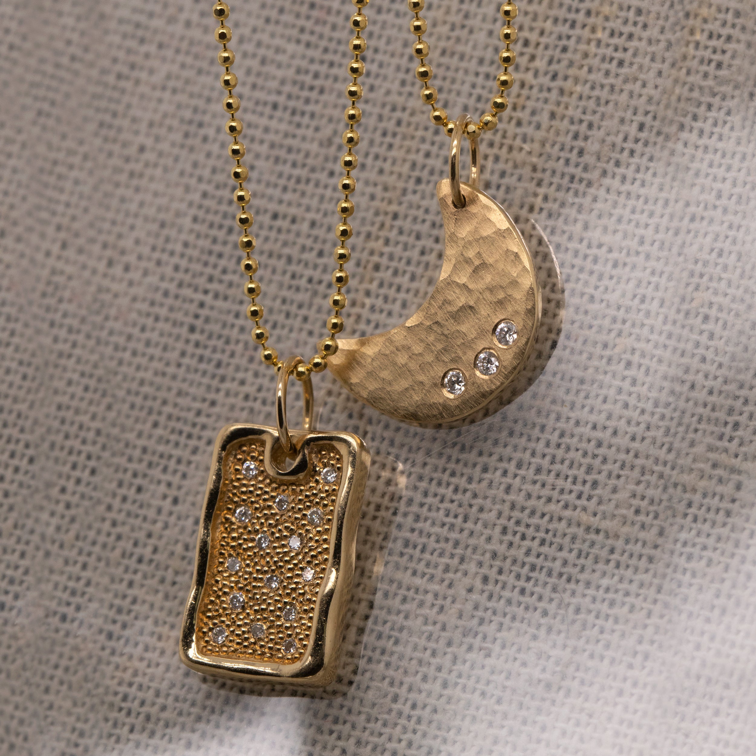 14k yellow gold necklace with a hammered crescent moon pendant featuring three small white diamonds hanging from diamond cut ball chain paired with rectangular 14k yellow gold dog tag with beaded texture and white diamonds on diamond cut ball chain on linen background