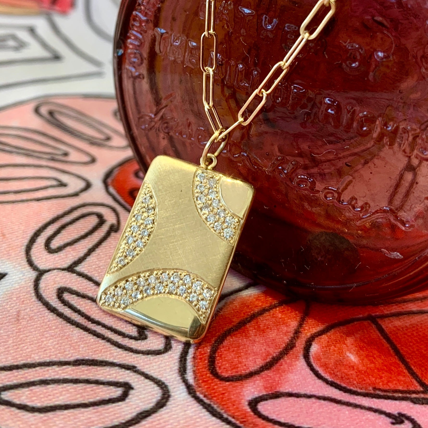 14k yellow gold rectangular pendant necklace with wavy diamond accents and a mix of shiny and satin finishes on link chain with colorful background