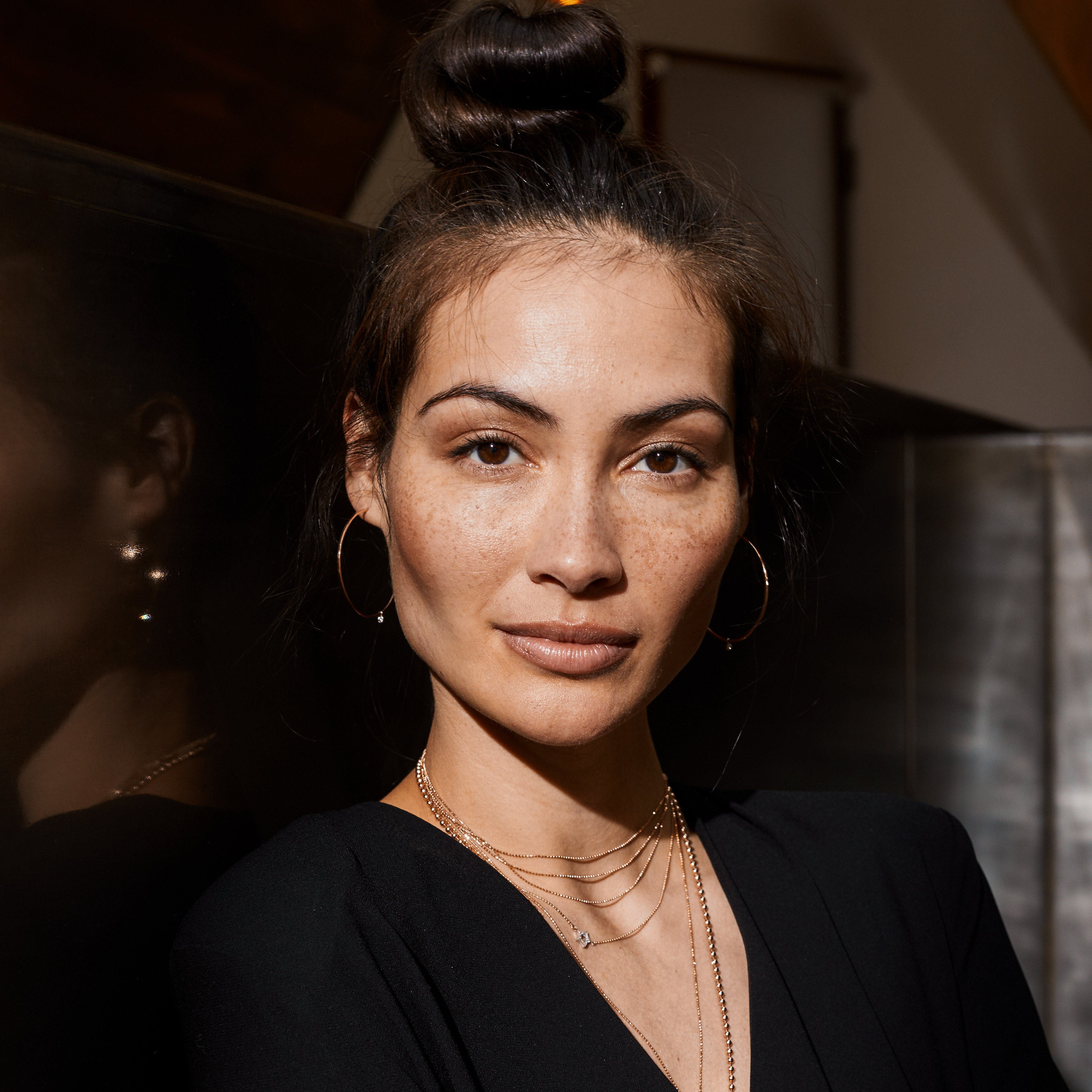 Woman wearing a black outfit with 14k gold jewelry, layered necklaces and large hoops with diamond accents standing indoors.
