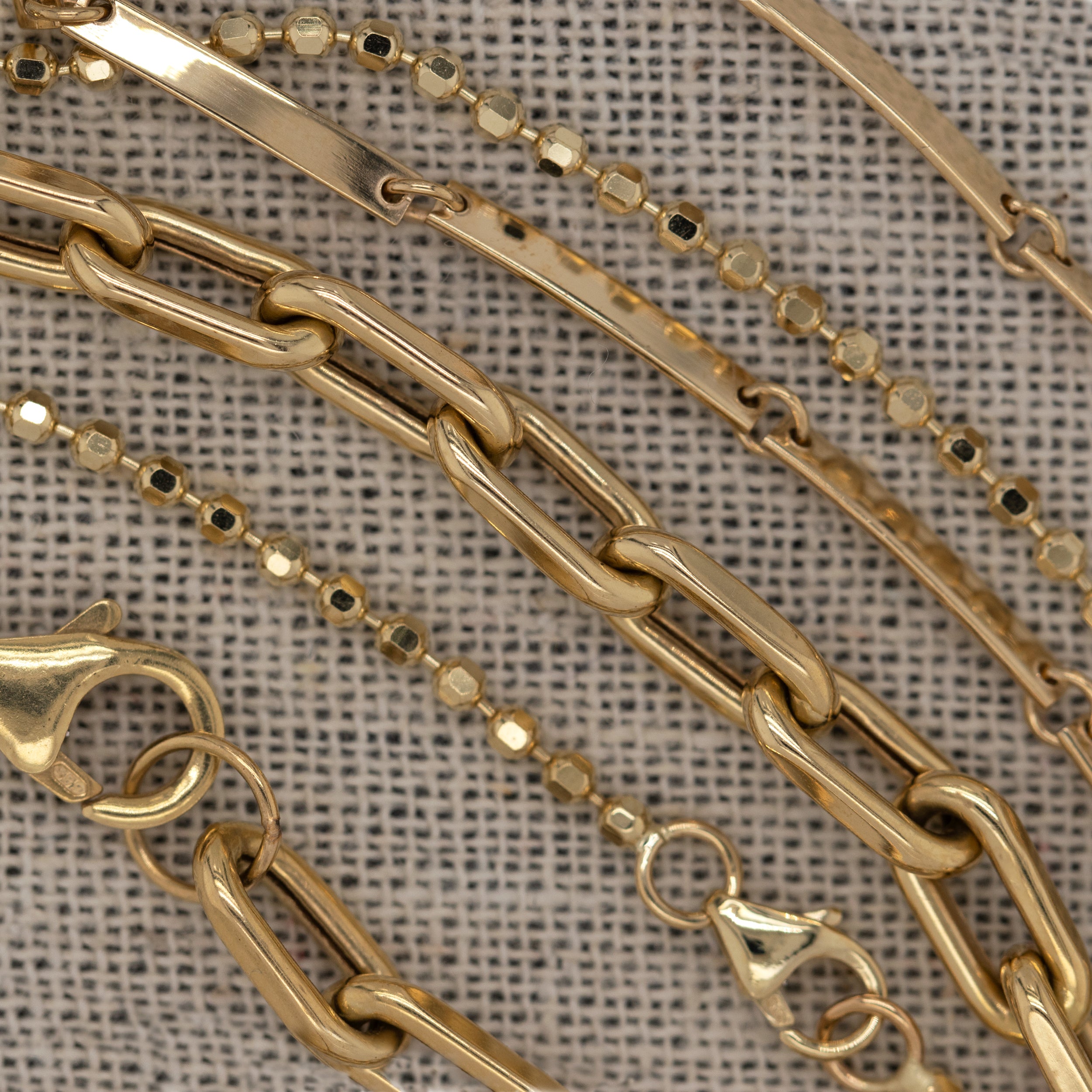 Variety of yellow gold bracelets detail shot faceted ball chain, flat link bar chain, and paperclip chains laying side by side on a textured beige fabric background