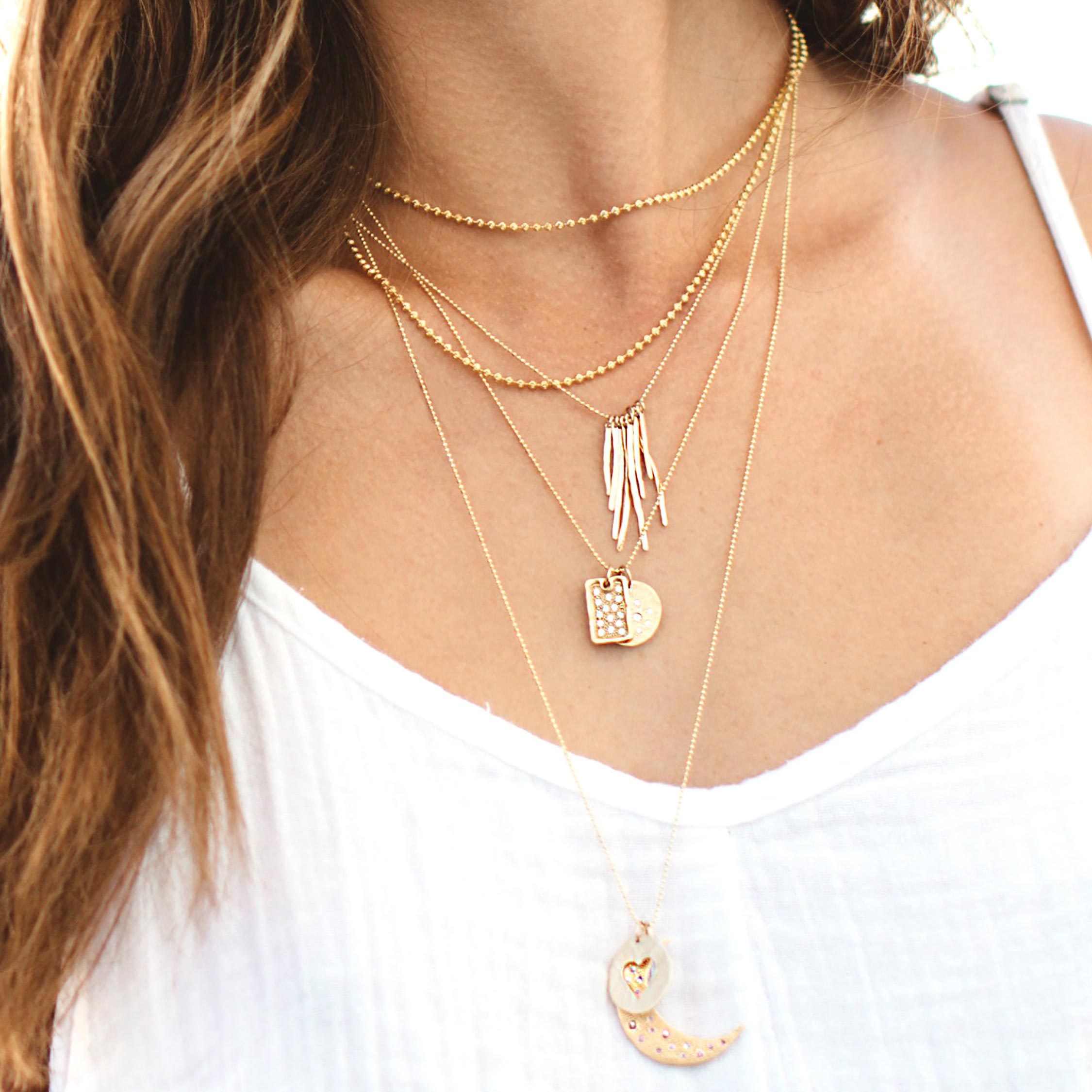 Close-up of a woman wearing multiple gold necklaces with layered necklaces including dog tag, heart and moon pendants on a lifestyle shot.
