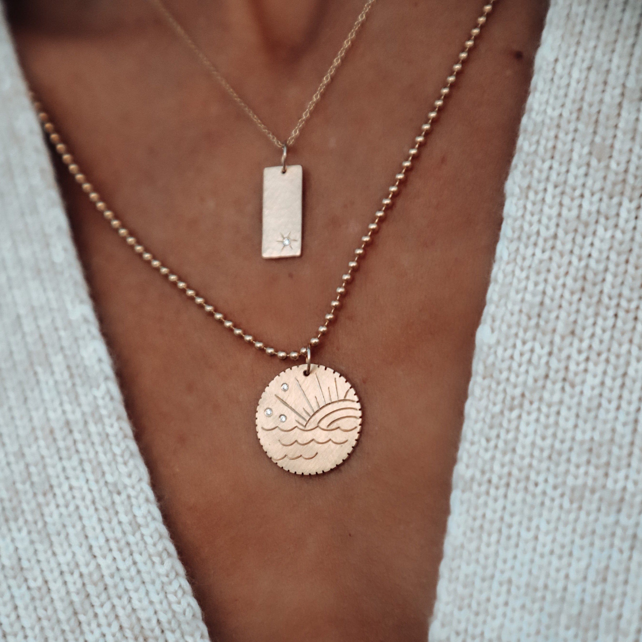 14k gold rectangular pendant with white diamond hand etched starburst on rolo chain paired with gone round charm with beach sunset engraving and white diamonds on ball chain in lifestyle shot on model