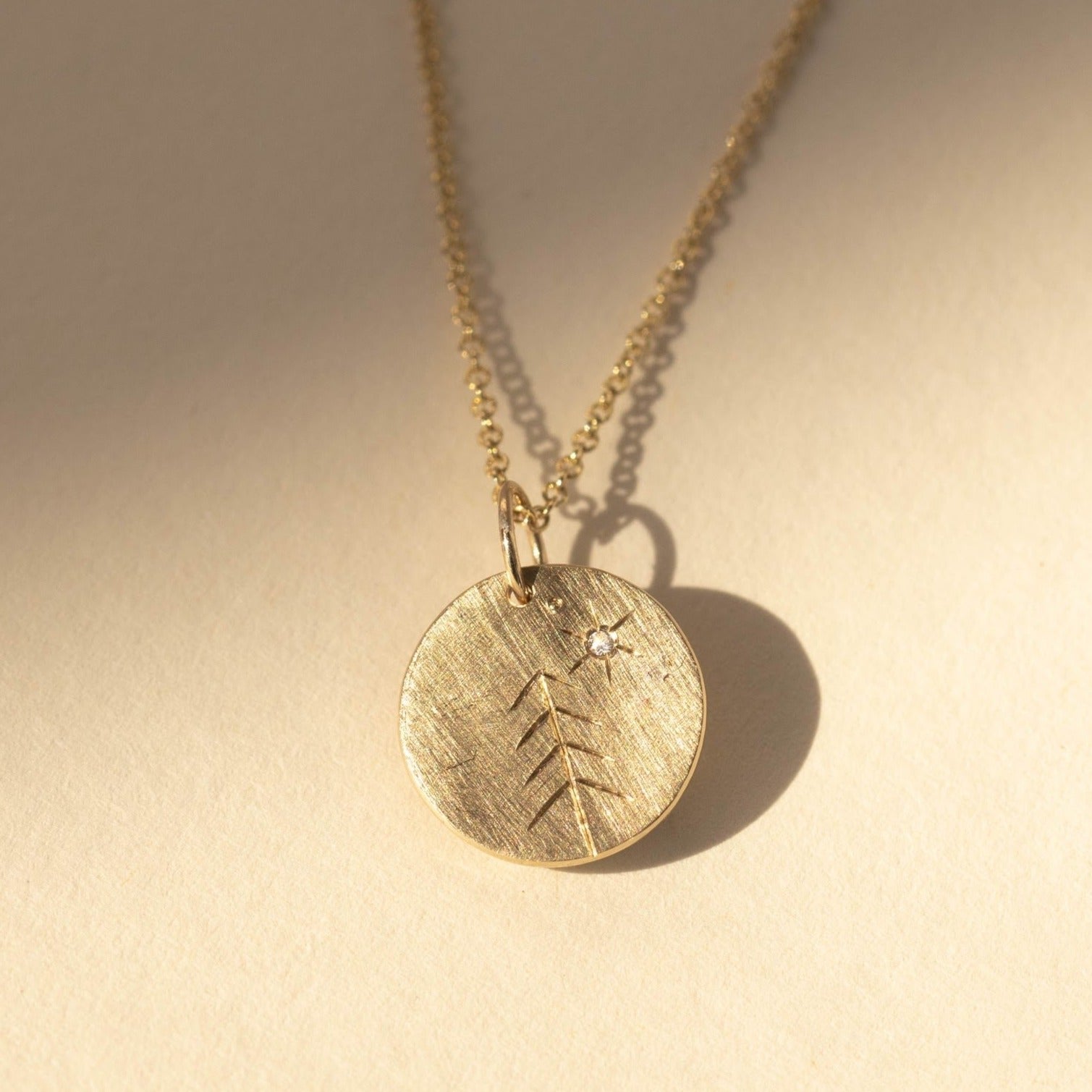 14k yellow gold round pendant featuring hand etched tree design and white diamond sunburst on rolo chain on a tan background