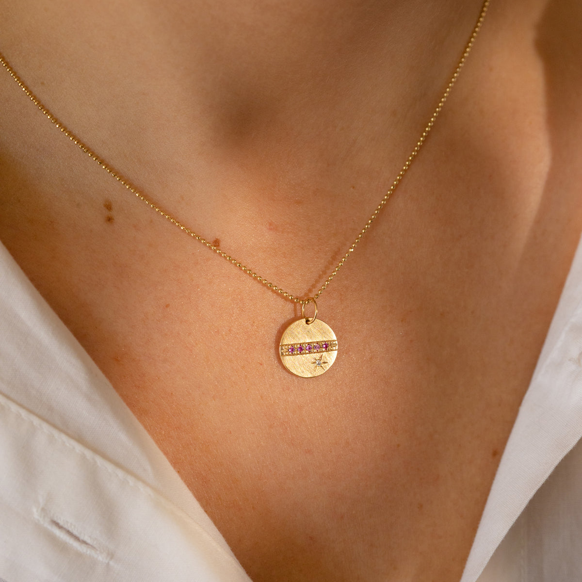 14k yellow gold round pendant featuring pink sapphires and a white diamond sunburst on diamond cut ball chain on model in lifestyle shot