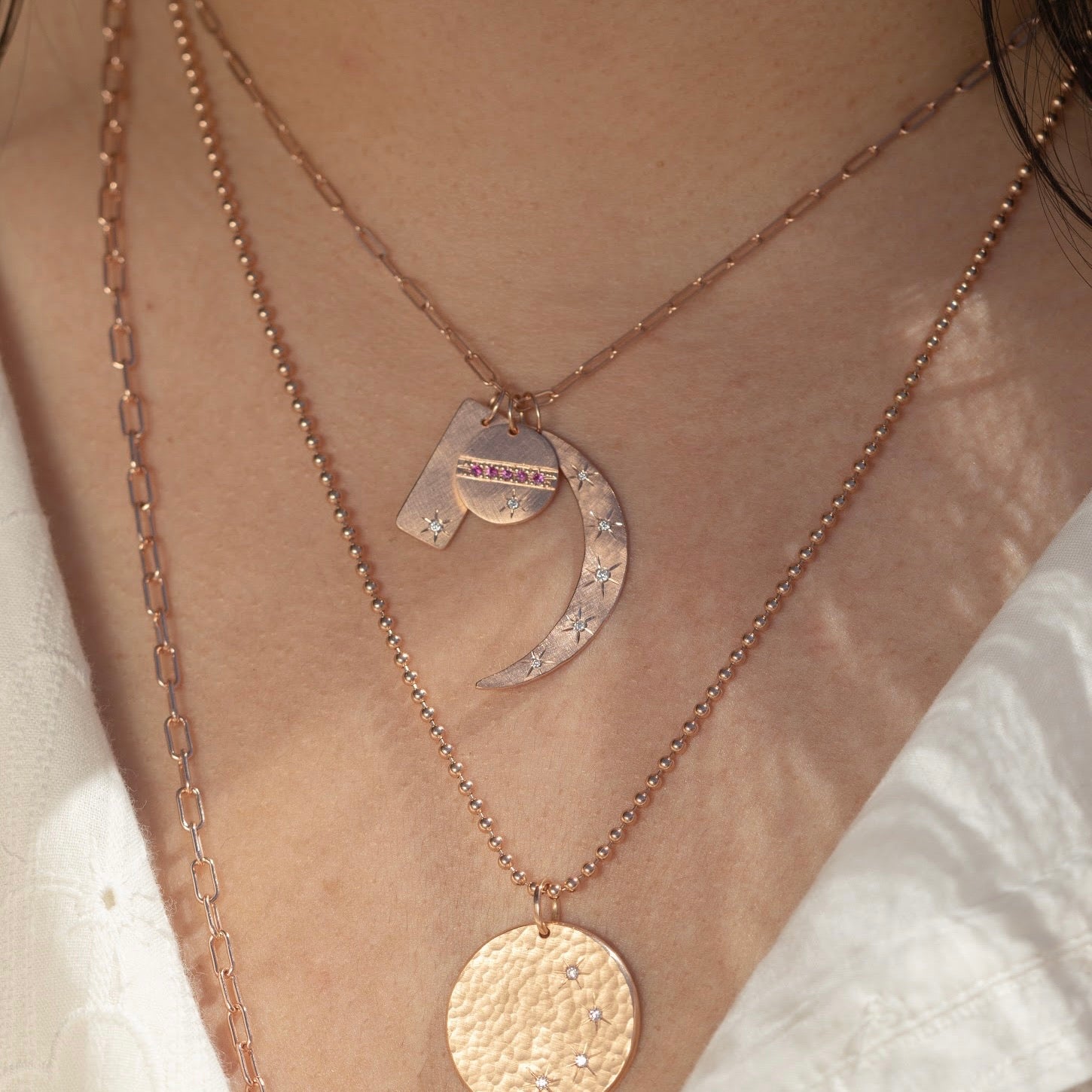 14k rose gold round pendant featuring pink sapphires and a white diamond sunburst on link chain paired with rectangular pendant and moon pendant stacked with round hammered pendant on model on lifestyle shot