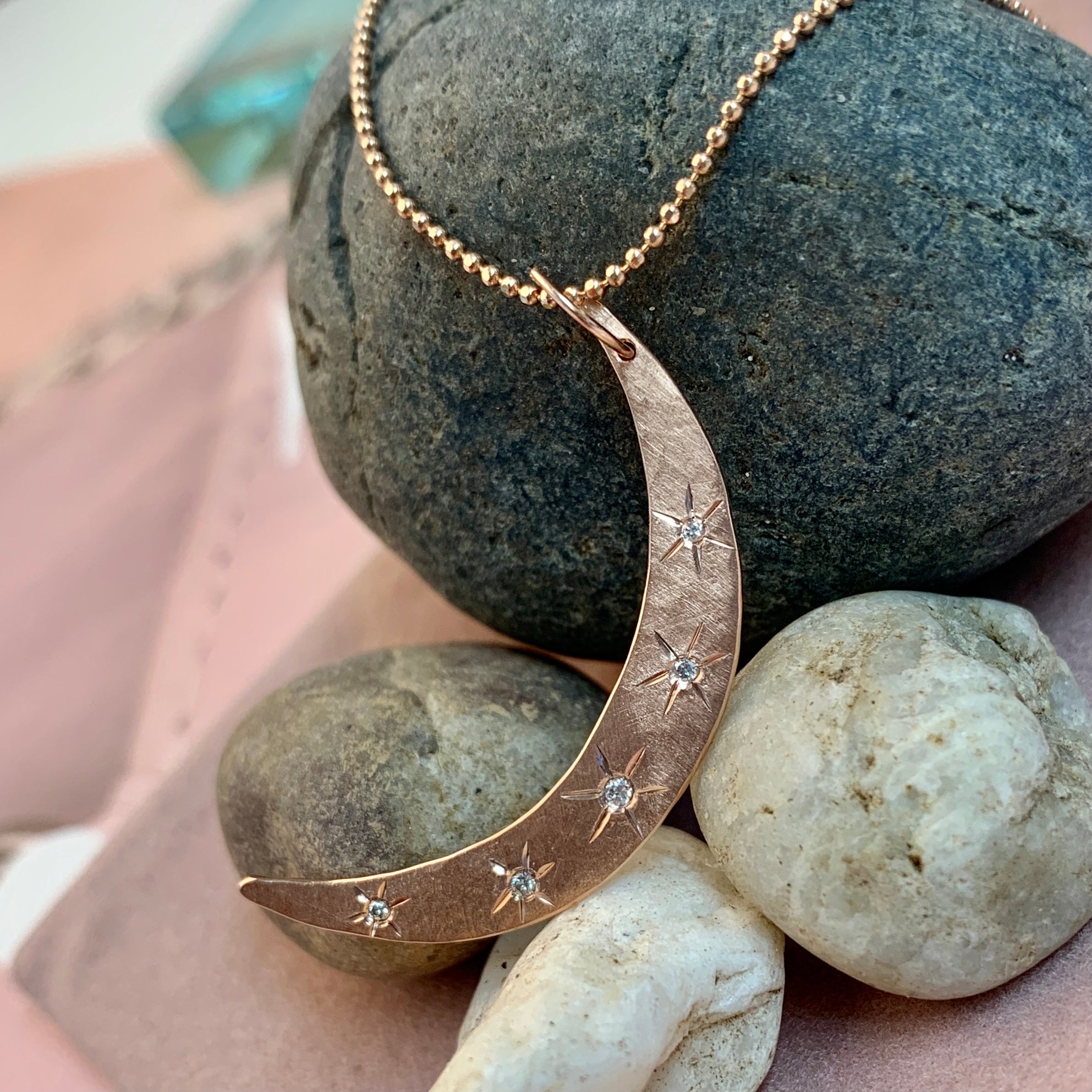 14k rose gold hammered crescent moon pendant with scattered white diamonds and hand etched starbursts on diamond cut ball chain on a rock background