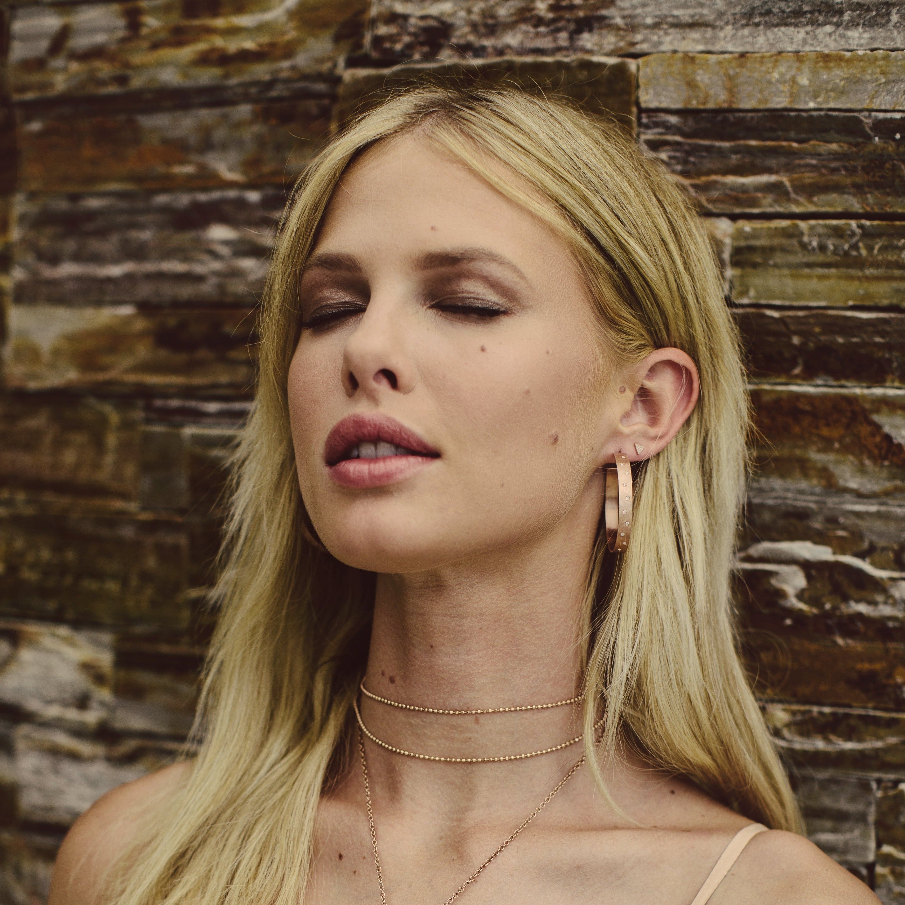 Woman with blonde hair wearing a pair of 14k gold large hoop earrings with a hammered, satin finish and scattered diamonds against a stone wall background