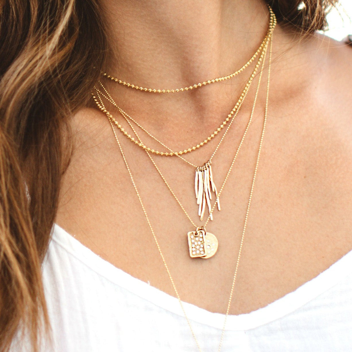 Close-up of a woman wearing multiple gold necklaces ball chain necklaces layered with heart and moon pendants, small dog tag, and fringe neck on a white background.aces by julez bryant