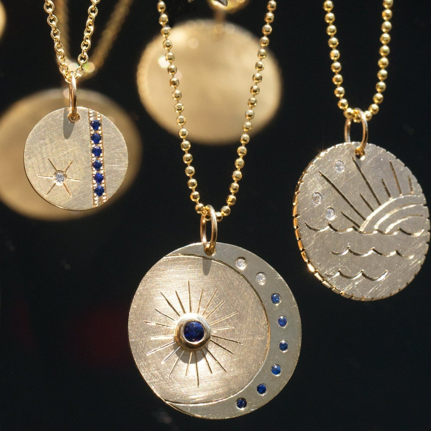 14k yellow gold round pendant featuring a shiny crescent moon and colored gemstones with hand etched sunburst stacked with two round pendants with hand etching and colored gemstones on a blurry background