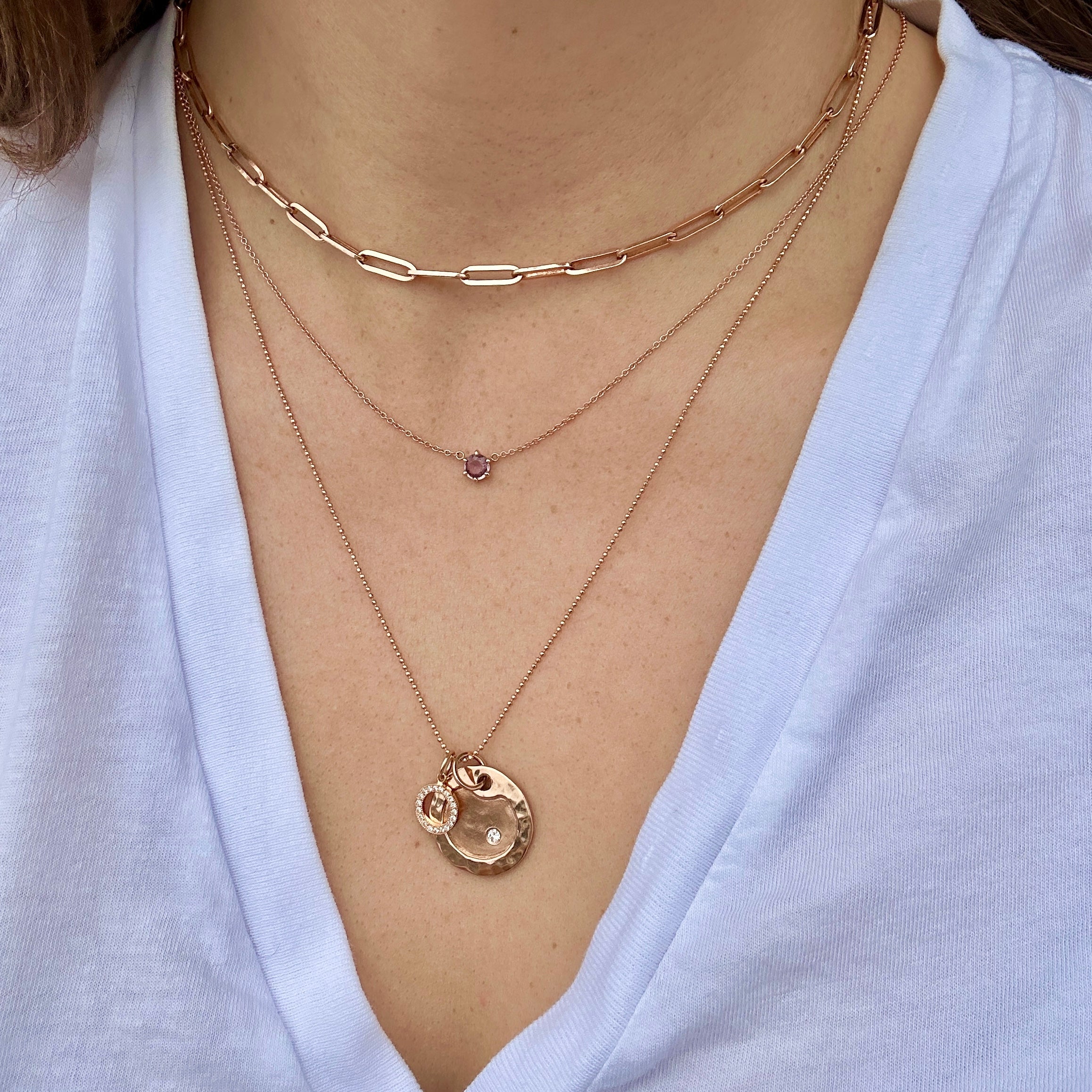 Close up of layered stack of 14k rose gold necklaces with full cut white diamond accents on a person wearing a light blue shirt