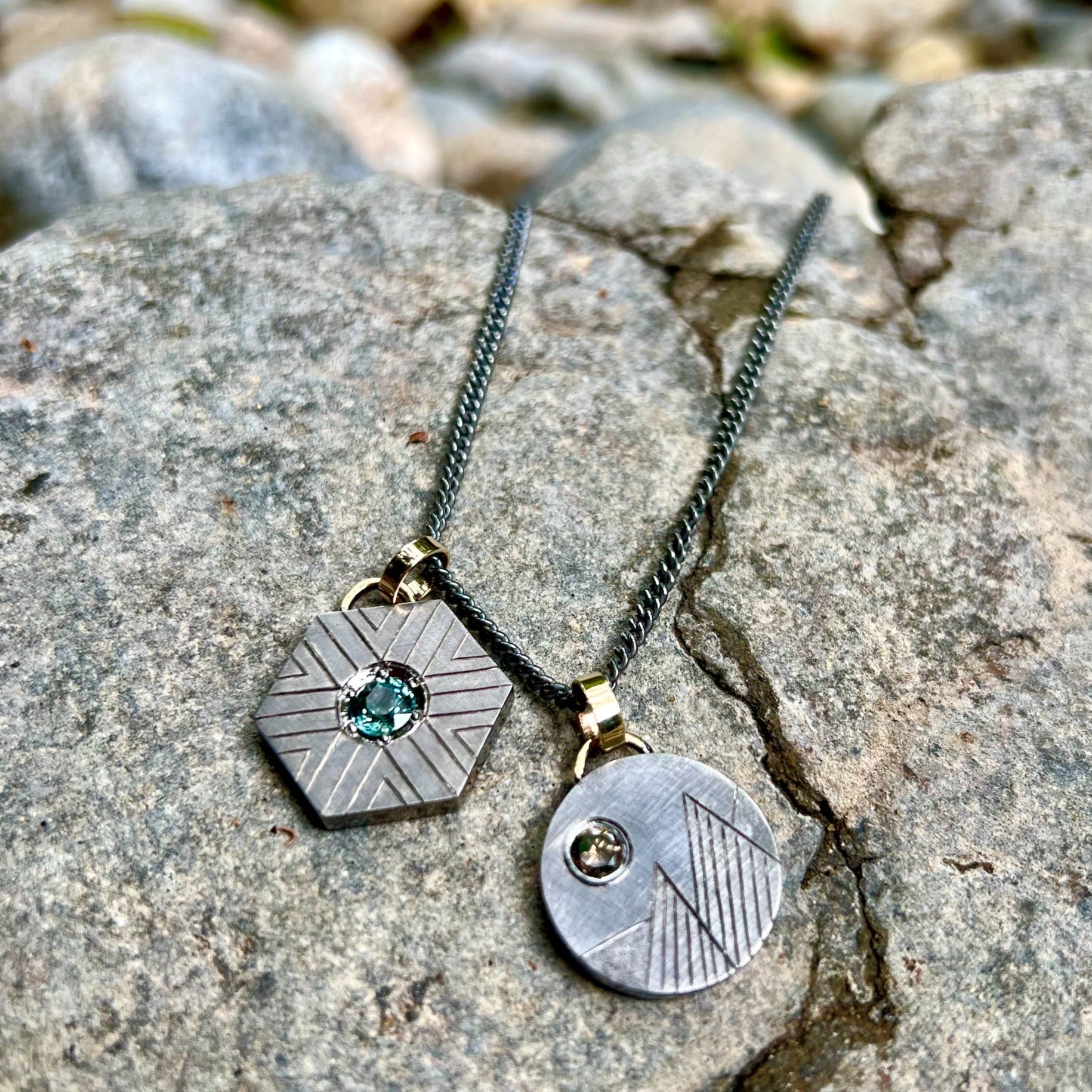 blackened sterling silver hexagonal pendant with geometric etching and a green sapphire with 14k yellow gold jump ring beside blackened sterling silver round pendant with a colored gemstone and etched mountains on rock background