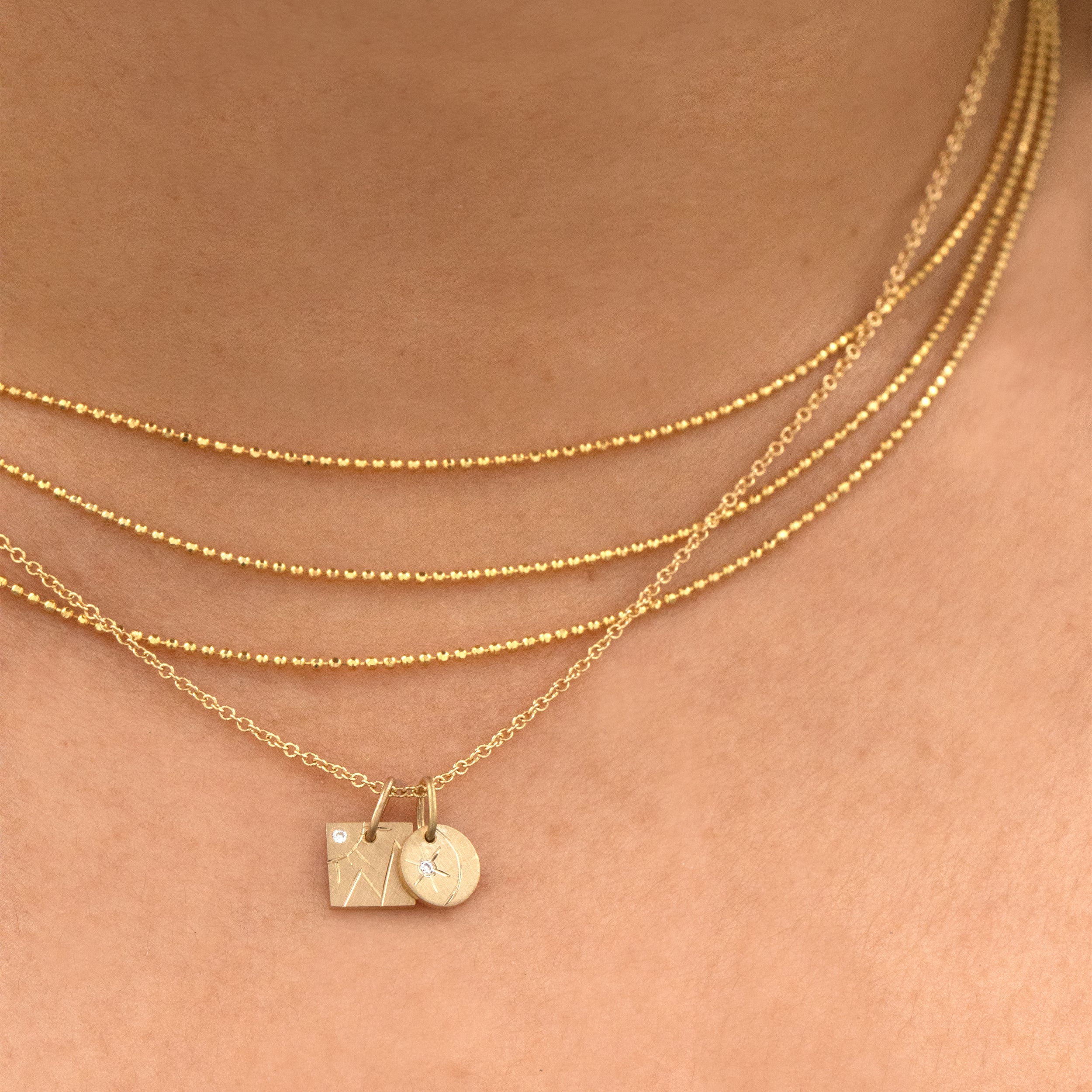 14k yellow gold round pendant featuring a diamond starburst with an etched crescent moon besides a square charm with mountain and sun etching with a white diamond stacked with the chai necklace on model in lifestyle shot