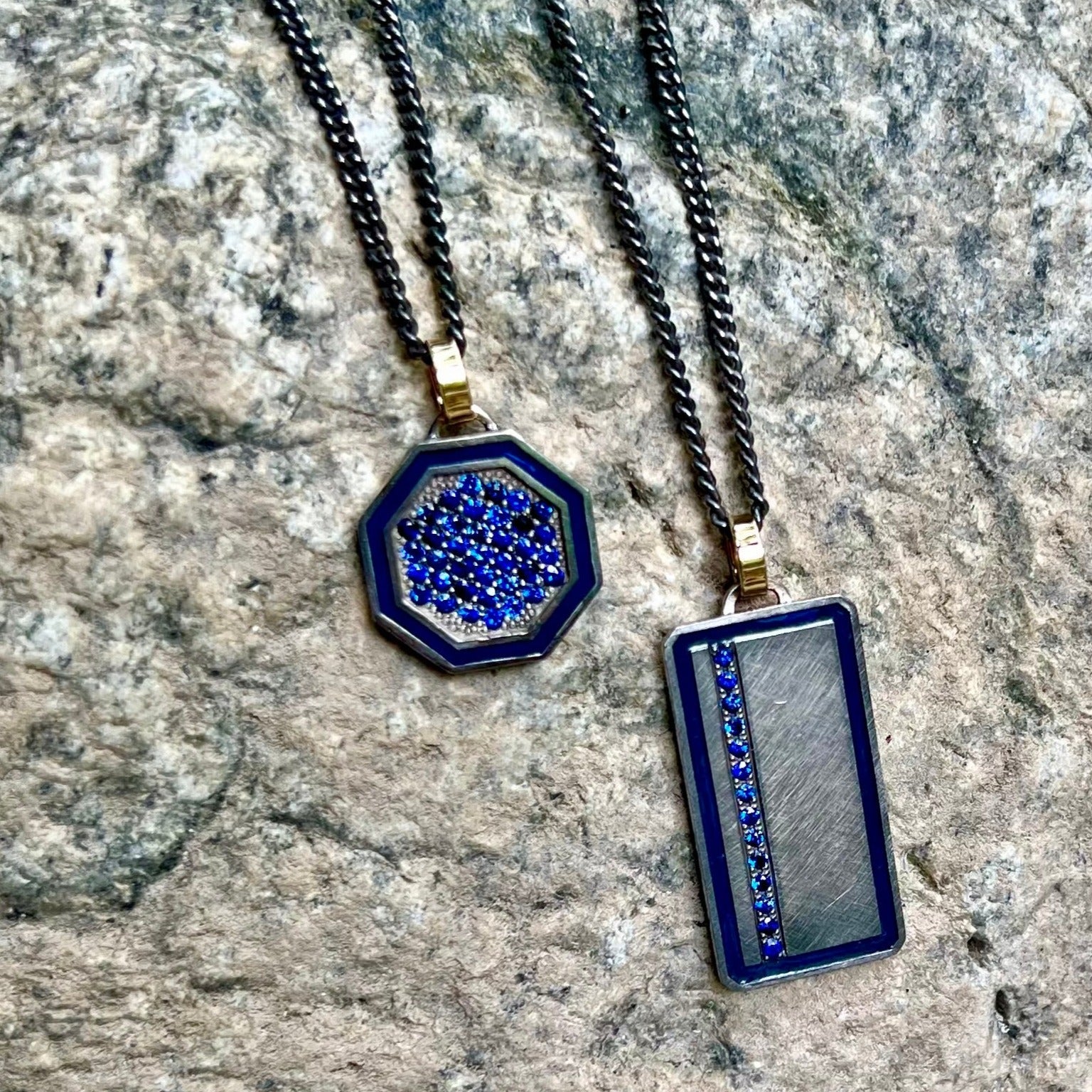 blackened sterling silver rectangular pendant with colored enamel and colored gemstone line with 14k yellow gold jump ring beside blackened sterling silver octagon pendant with colored enamel border and colored gemstones with 14k yellow gold jump ring on rock background