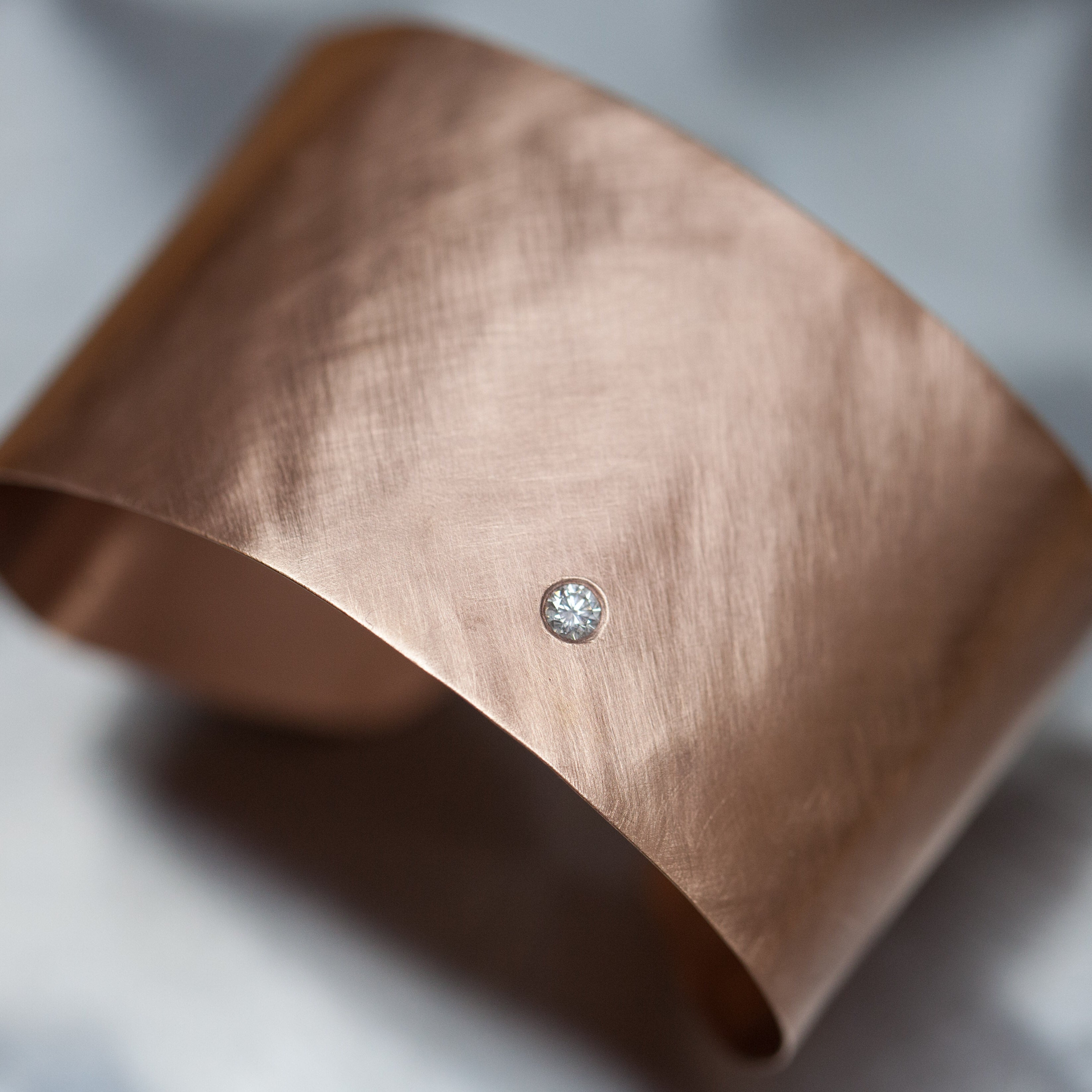 Rose gold extra wide cuff bracelet with a single white diamond accent, brushed texture finish shown on a gray background