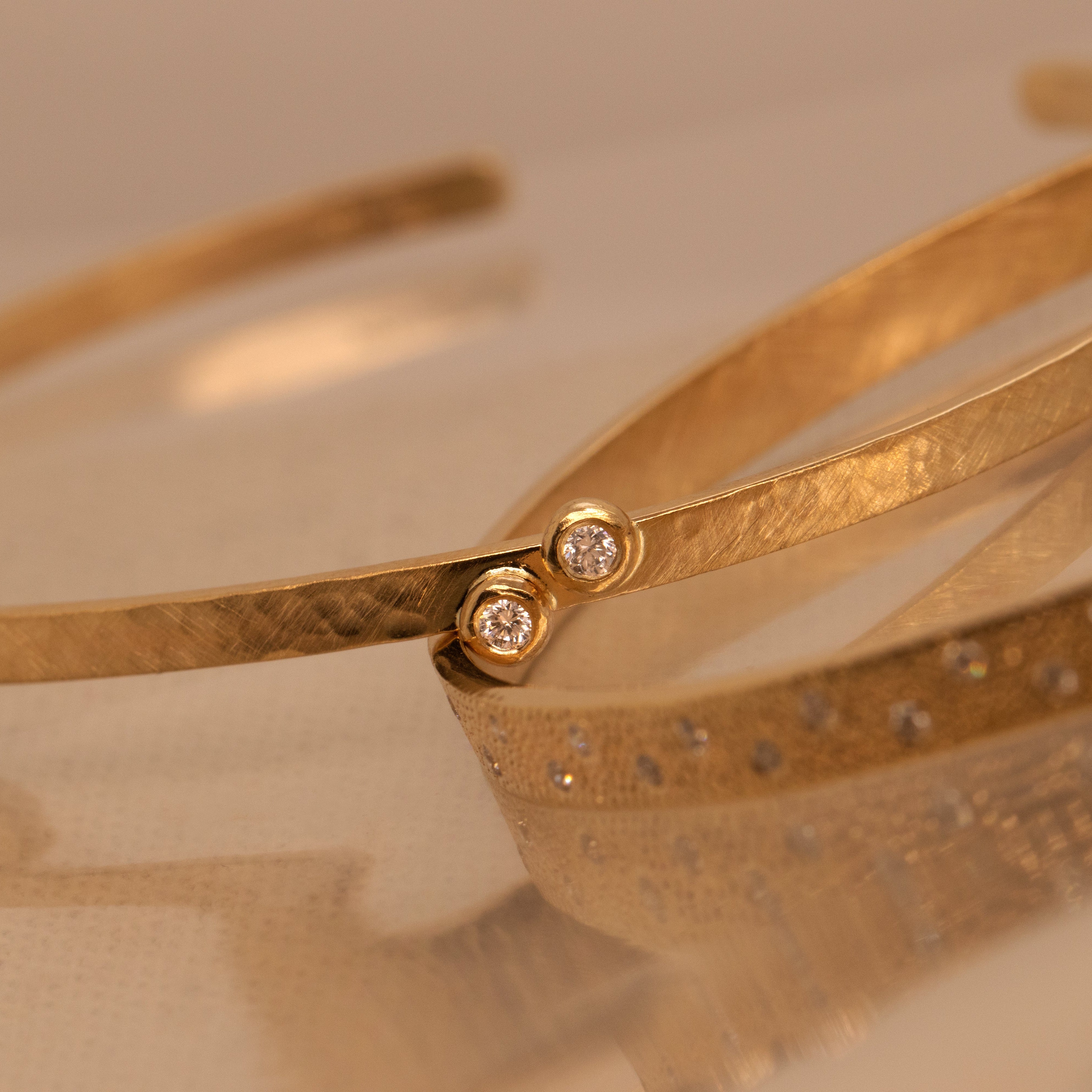 Gold cuff bracelets featuring two bezel set white diamond on a thin hammered satin finish cuff on a reflective surface