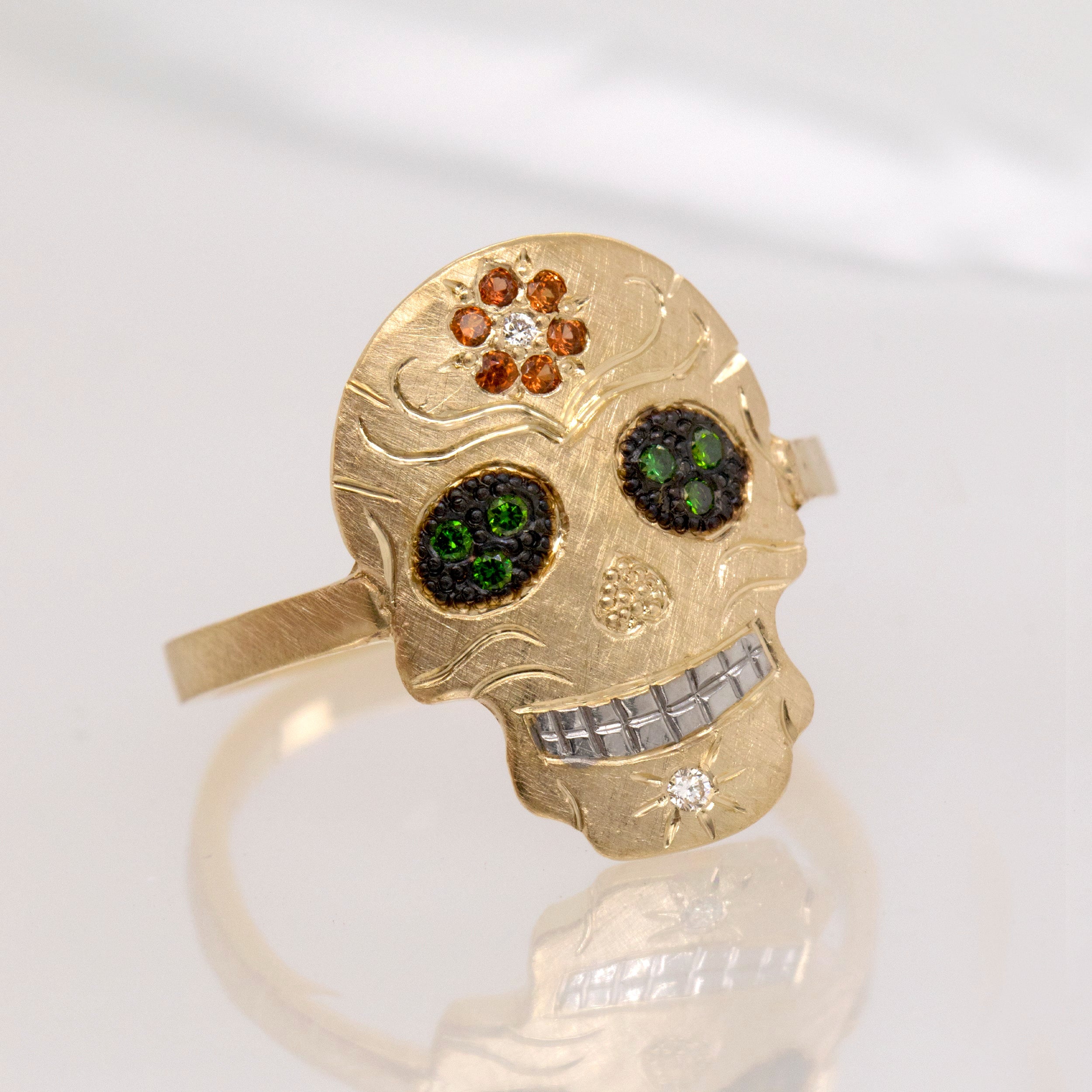 Gold skull-shaped ring with gemstones on a white background. Pave set orangesapphire flower on forehead, pave set eyes and diamond on chin. Fine engraving and milgrain detail. 3/4 view one white reflective background