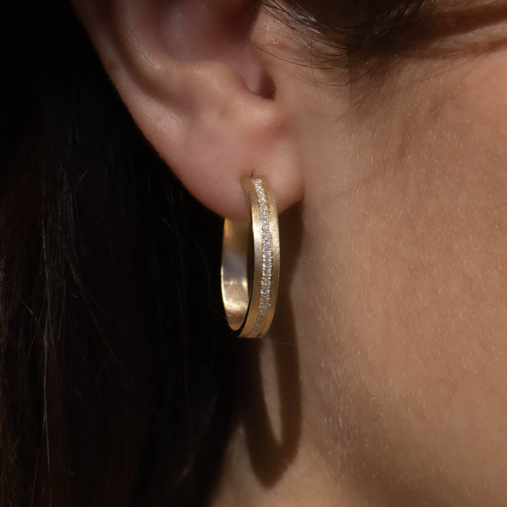 close up of womans ear wearing a pair of 14k yellow gold diamond hoop earrings with a satin finish and a continuous strip of paved set white diamonds blurred background