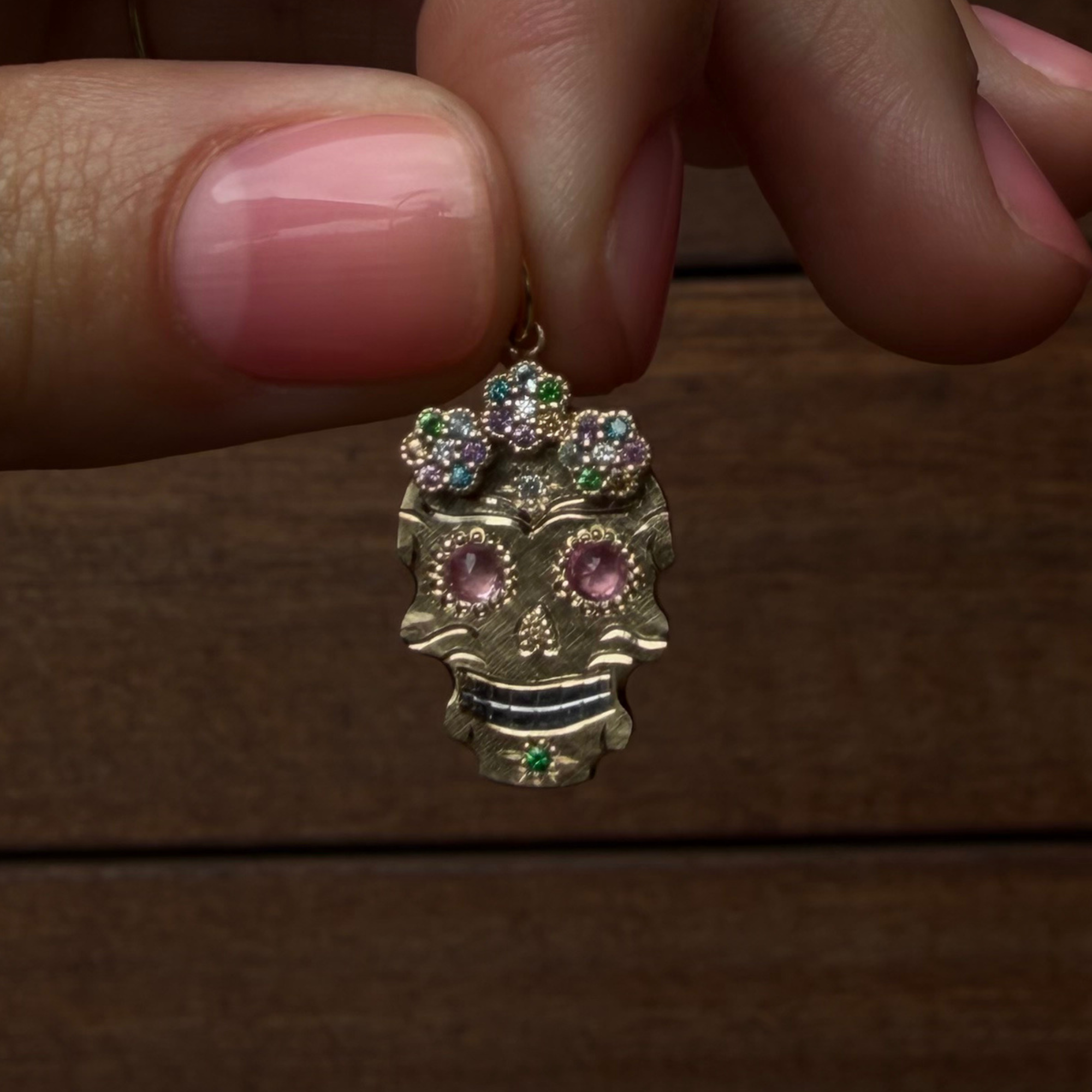A close-up of a hand holding a gold sugar skull pendant with pink sapphire eyes and multicolor rhinestone floral halo against a dark wood background.