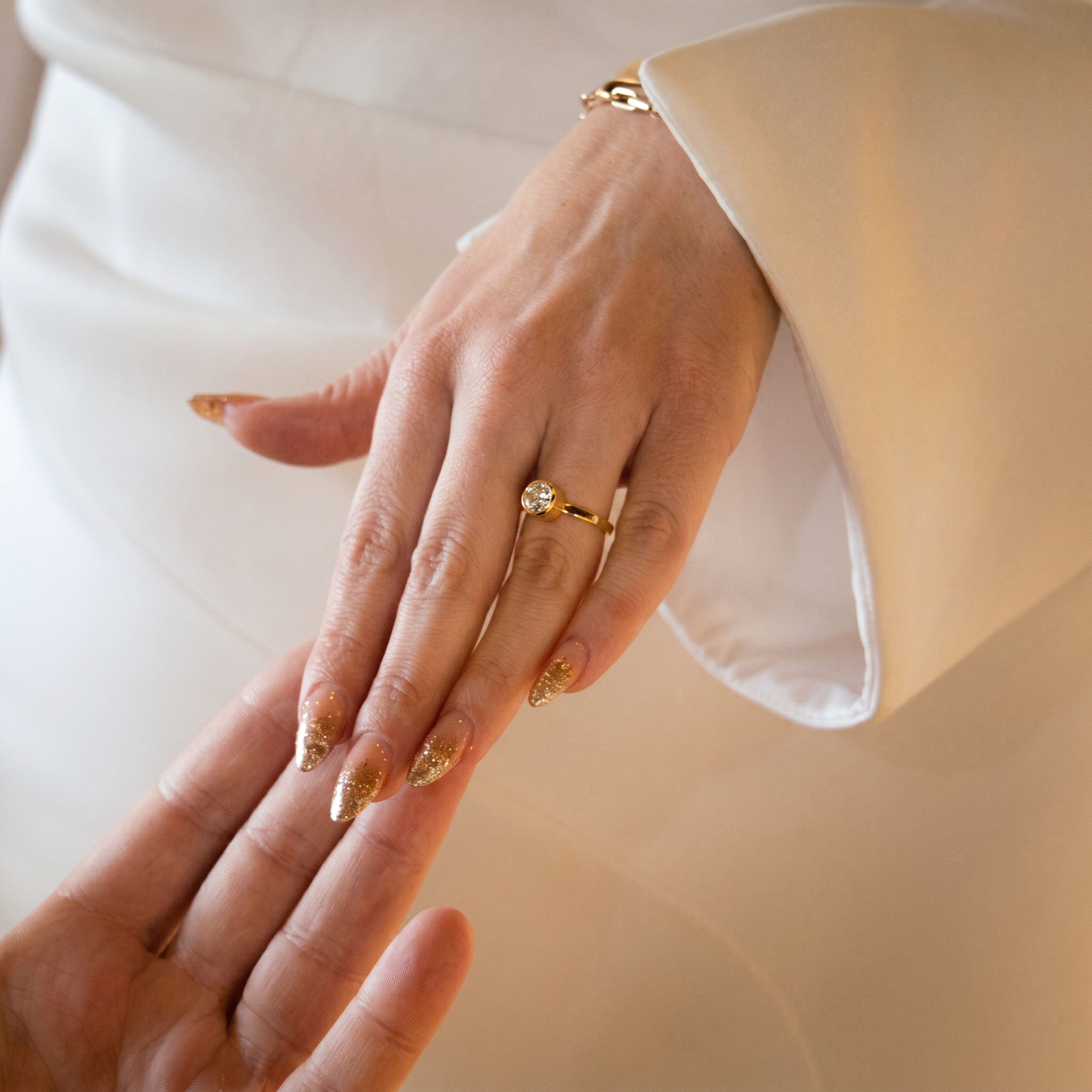 Round brilliant bezel solitaire engagement ring worn on a womans hand with gold nail polish wearing white dress. Close up of two hands touching.