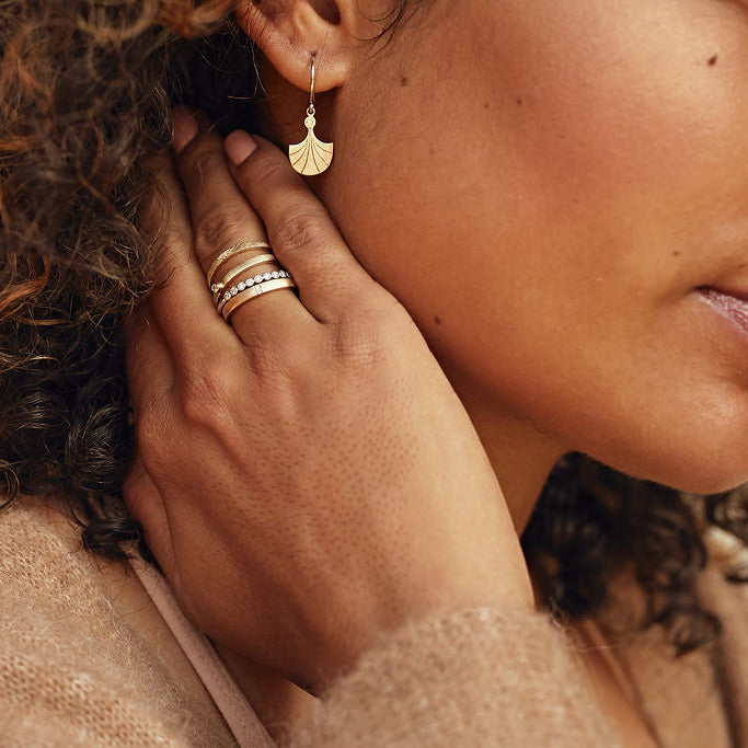 Model wearing dainty fan shaped drop earrings with tiny white accent on french hoop and stack of thin bands blurred background