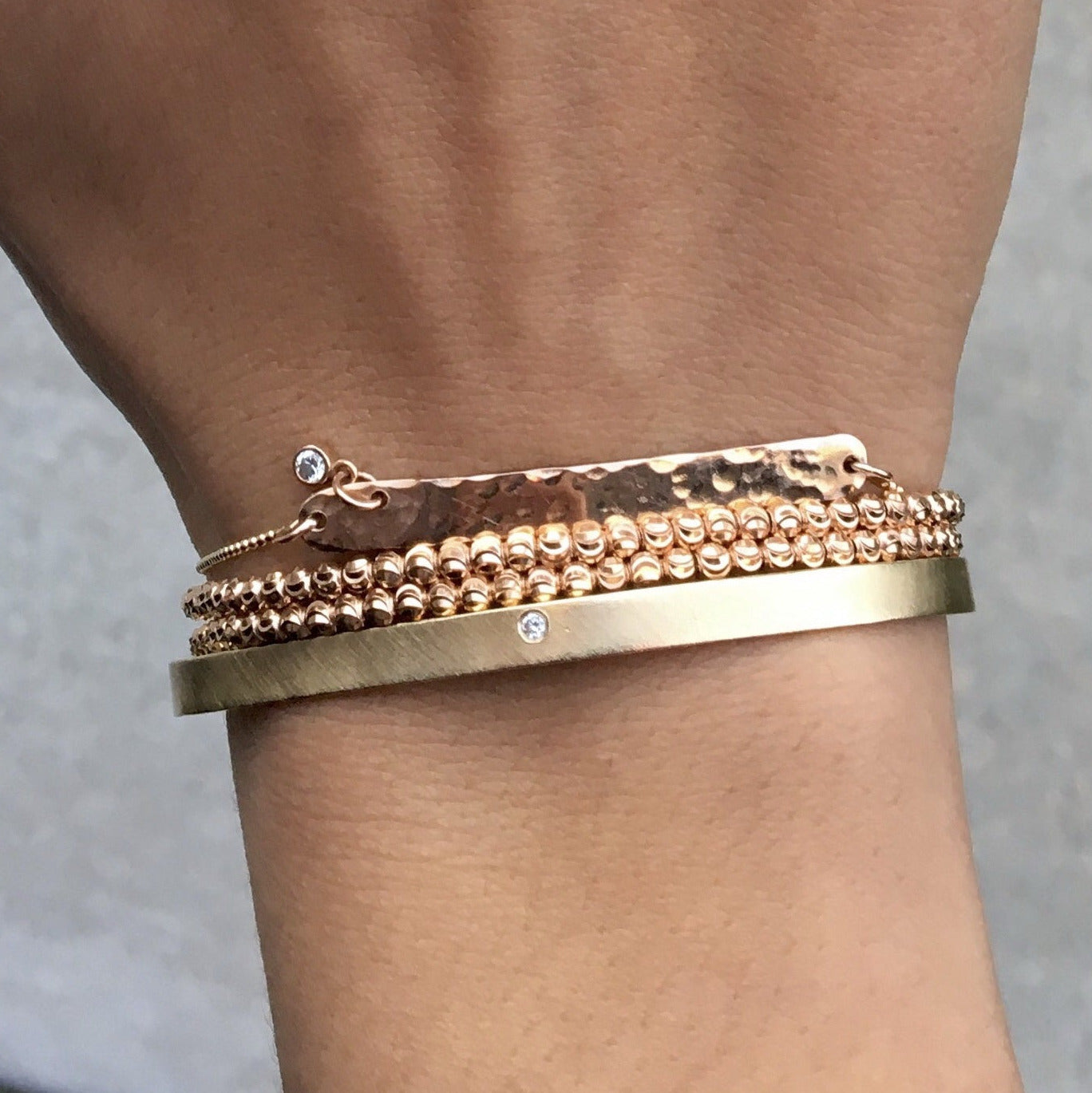 Close-up of a wrist wearing multiple gold bracelets, cuff, faceted beaded, double wrap and bar chain on a neutral background