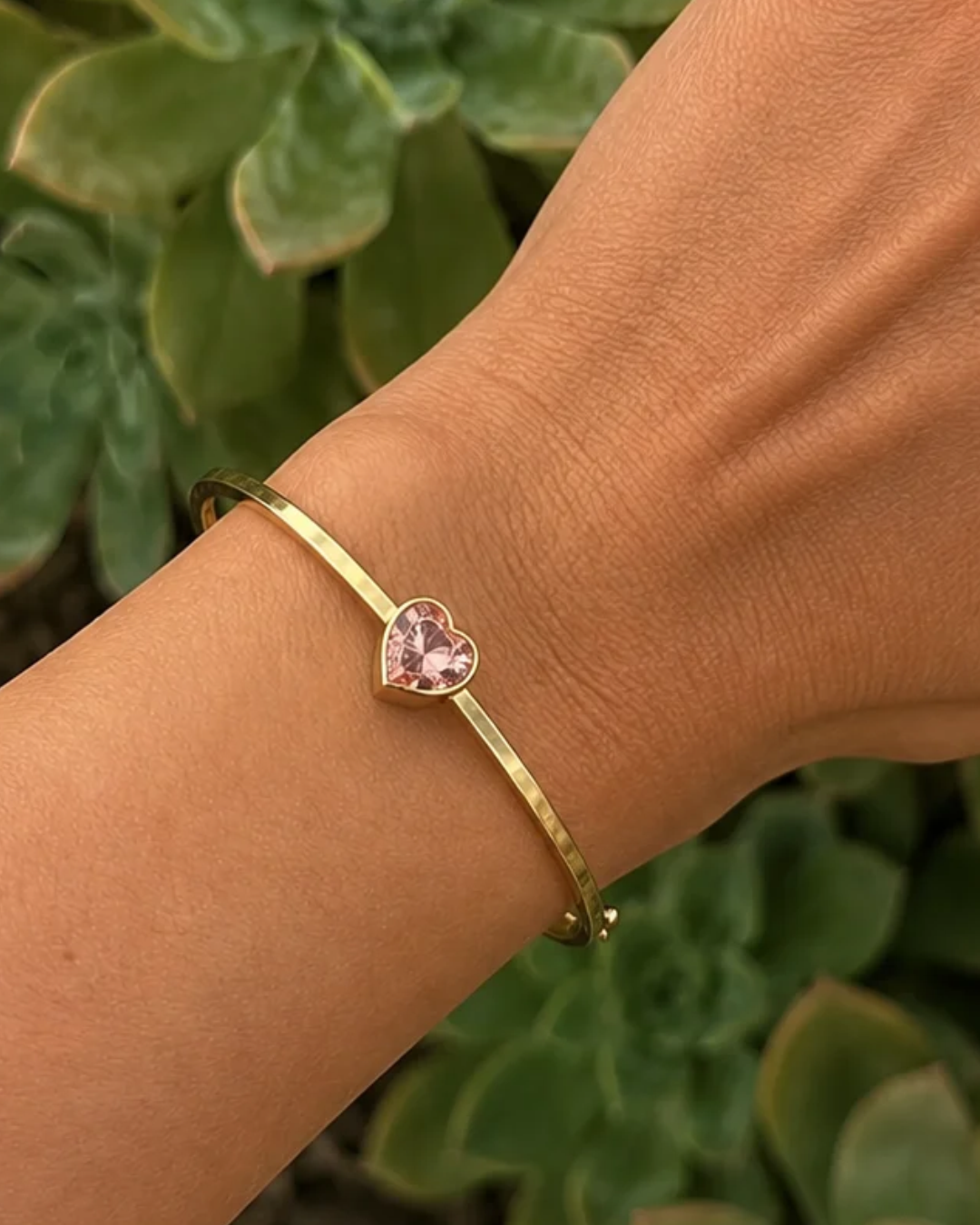 A gold open cuff bracelet with a heart-shaped pink gemstone, worn on a person's wrist with a blurred green succulent background.