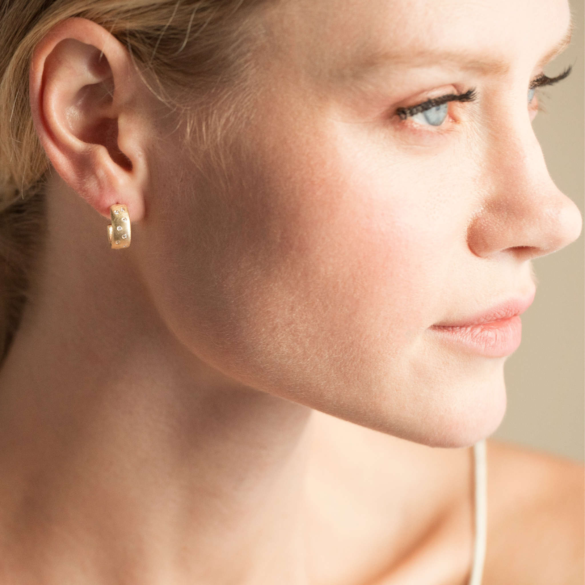 Model wearing small 14k yellow gold huggie hoop earrings with scattered white diamonds close up of face with blurred background.
