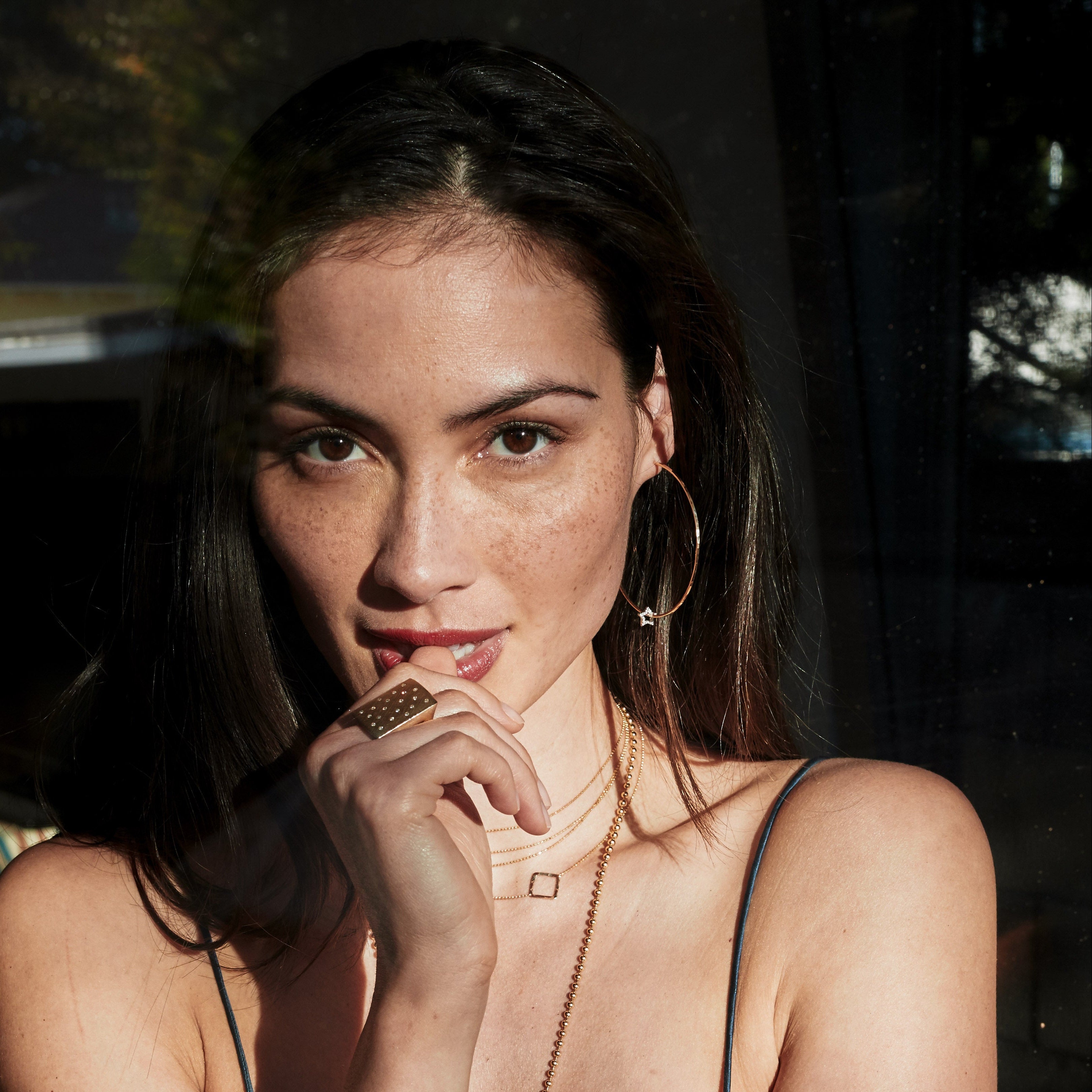 Woman wearing a blue top and gold jewelry with white diamond accents including large hoop earrings and a block ring in a dimly lit setting