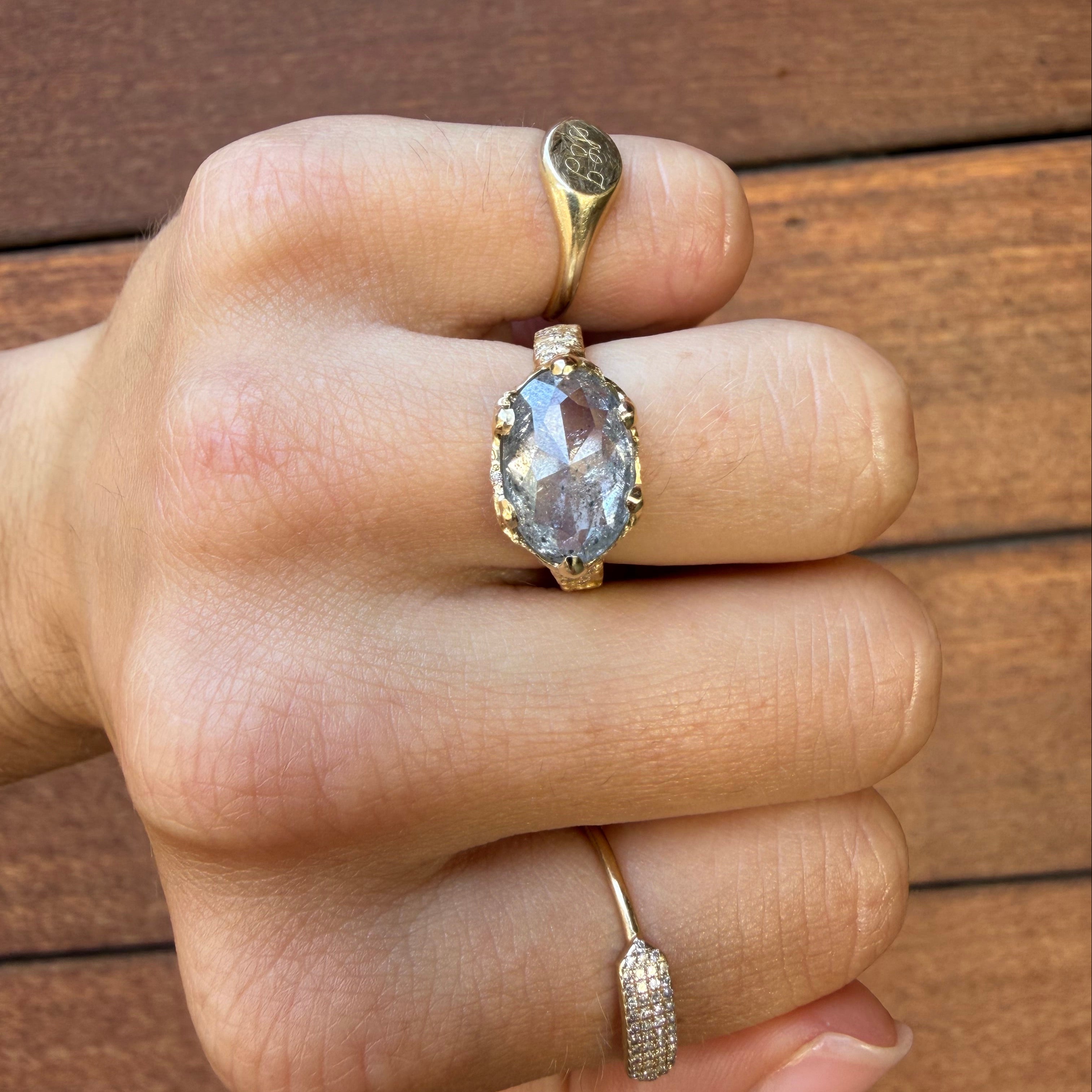 A close-up of a hand wearing multiple gold rings, including a prominent oval-cut light blue gemstone ring on the middle finger, set against a brown wooden background.