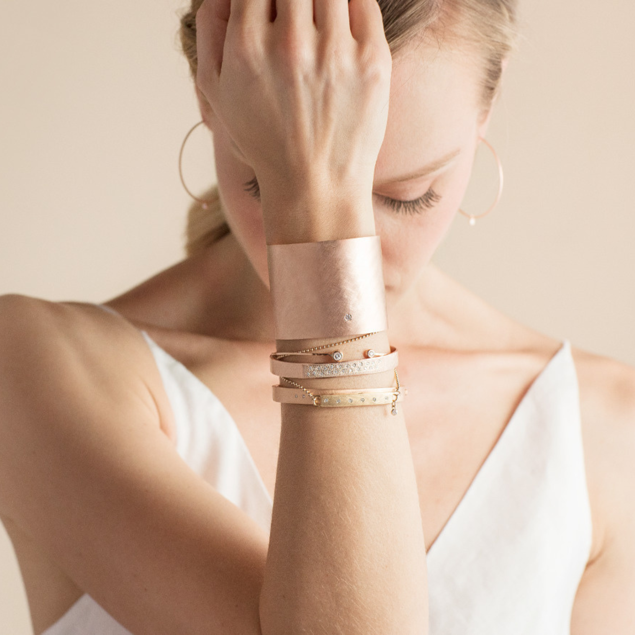 Model wearing stacked bracelets in rose and yellow gold. Double wrap bar bracelet bracelet paired with Statement hammered cuff layered with medium weight cuffs and and thin paddle bracelet white diamond accents hand to face with blush background.
