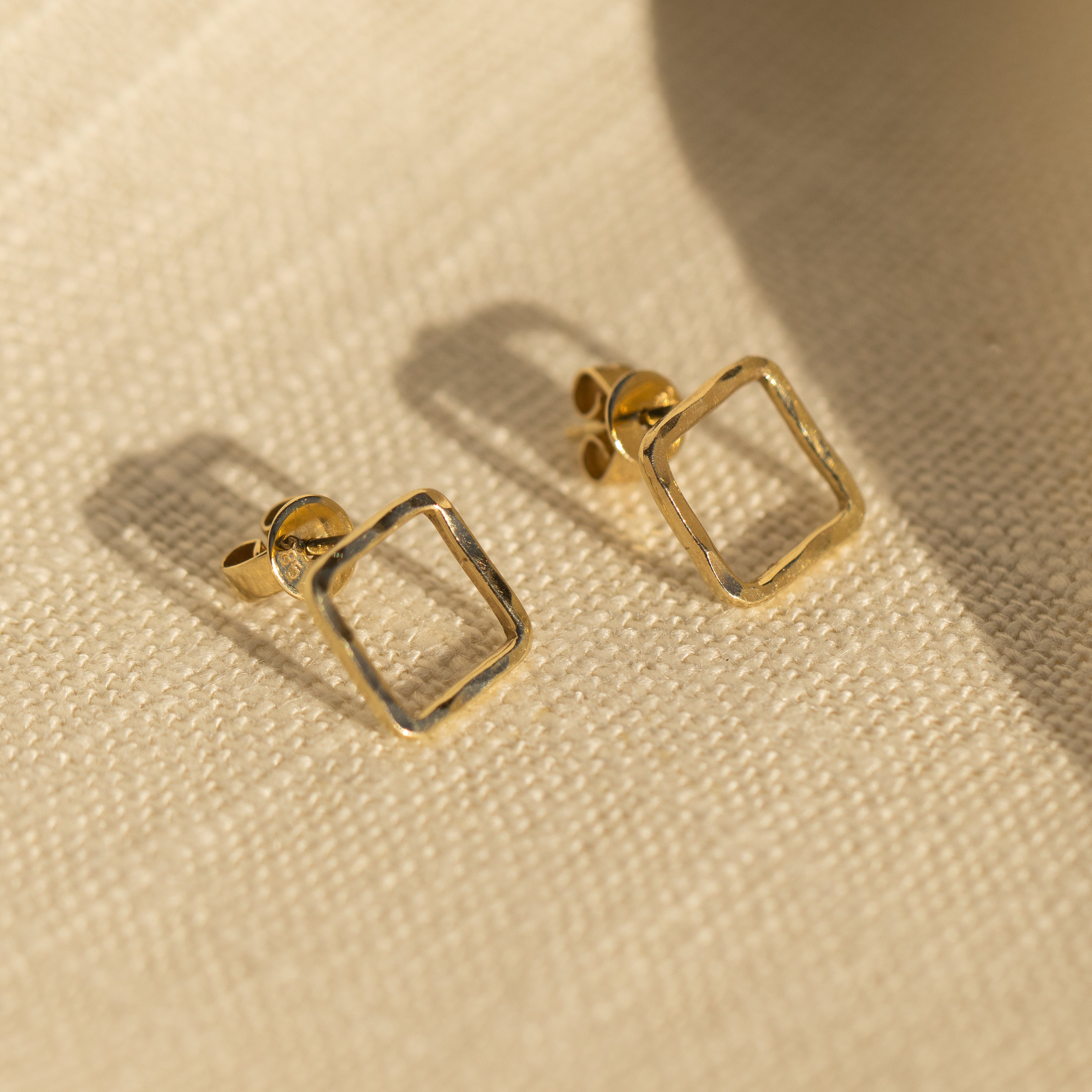 Lifestyle shot of 14k gold open square shaped post earrings with hammered texture 3/4 view laying on linen background
