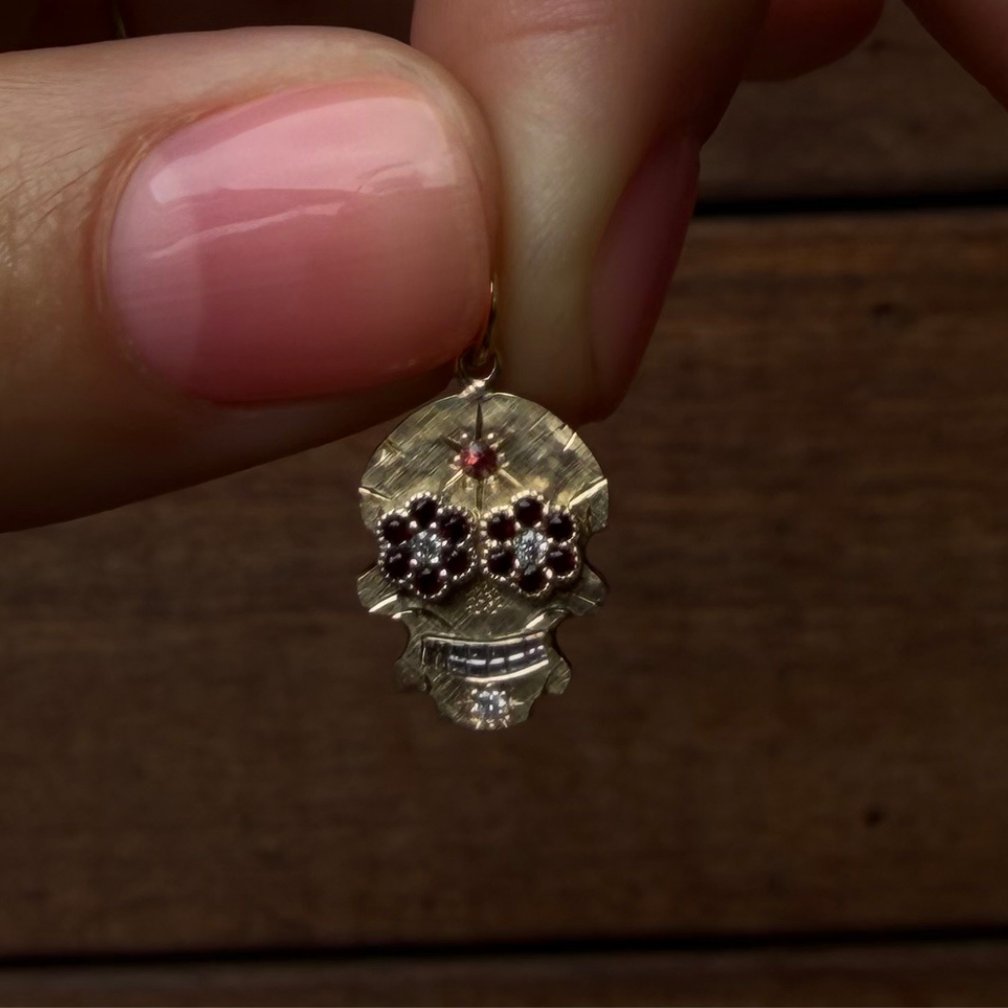 A close-up of a hand with pink manicured nails holding a small gold skull pendant with dark and clear stones, set against a dark wooden background.