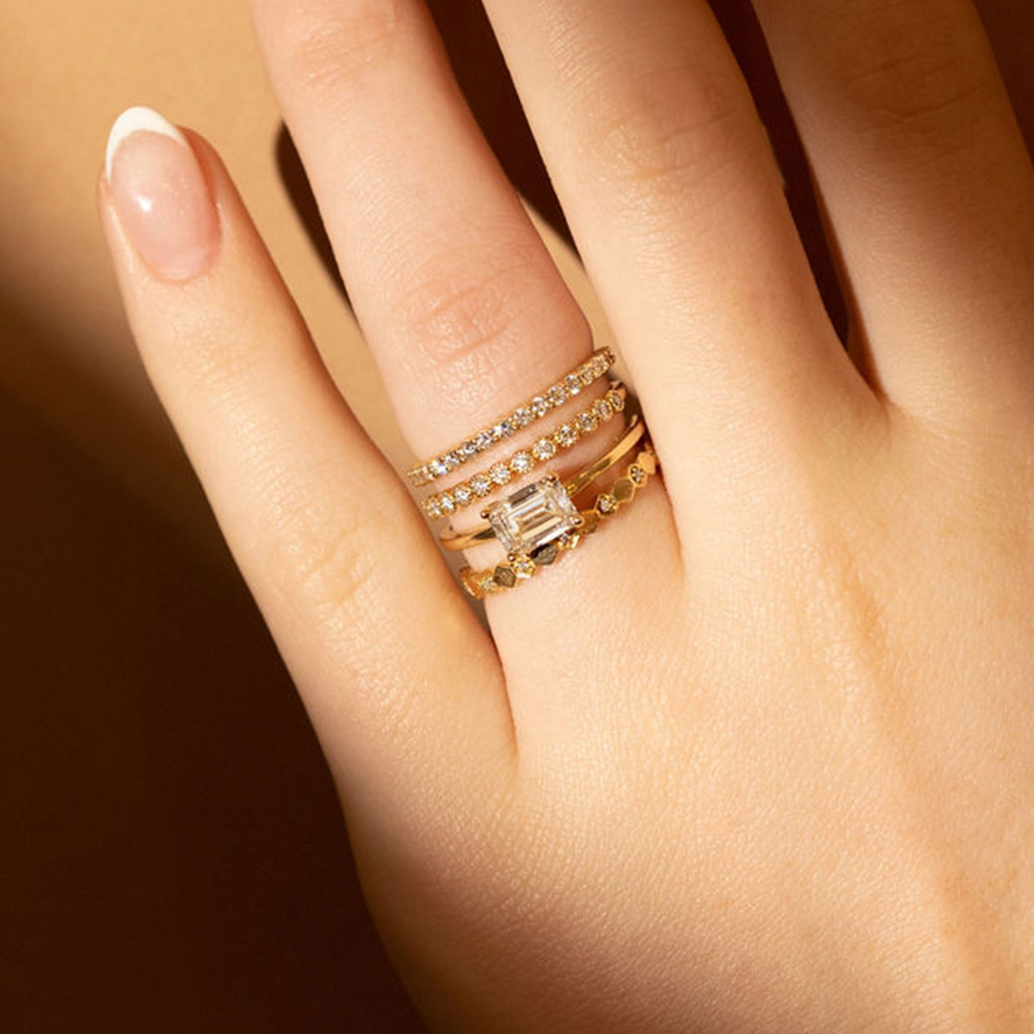 Close-up of a hand wearing four gold rings with white diamonds featuring eternity bands, hexagon band and emarald shaped solitaire ring on a warm-toned background