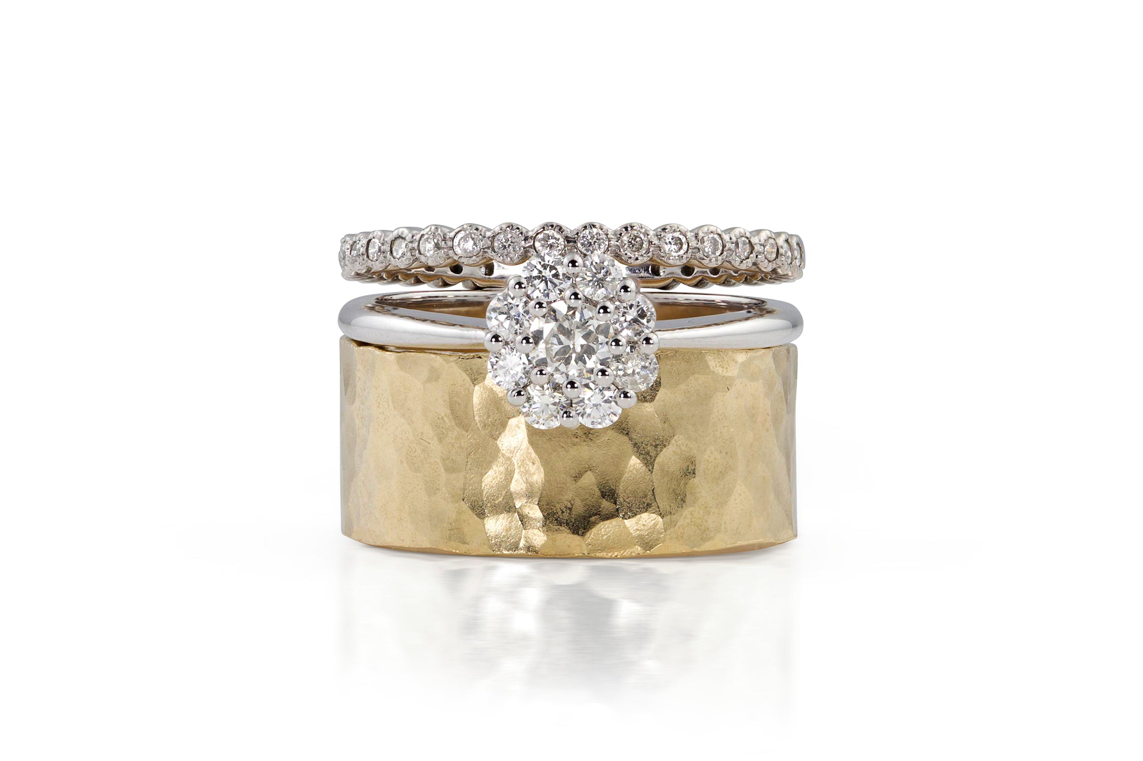 Three diamond-studded rings in white and yellow gold with milgrain, cluster and wide hammerd textured band on a white background