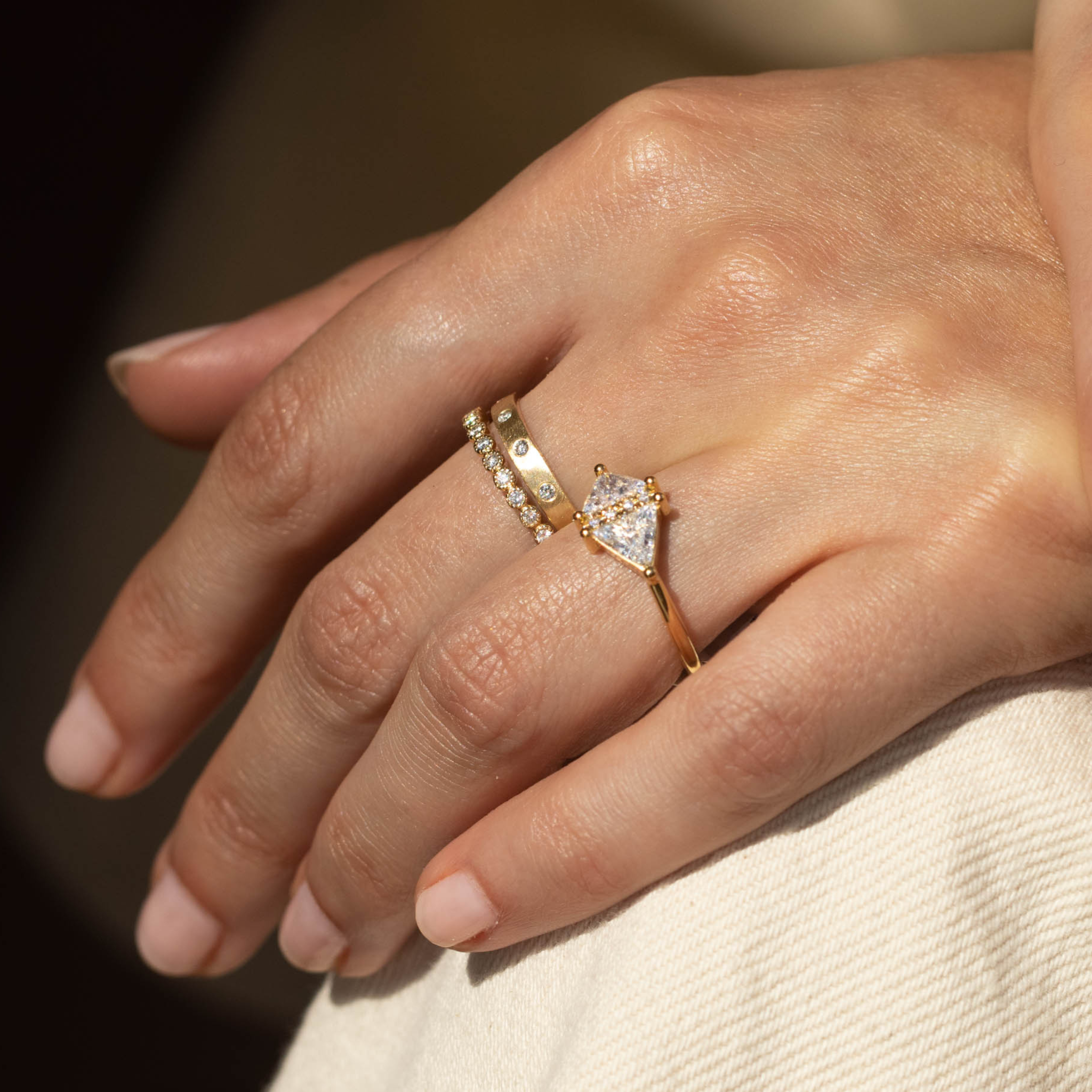 Close-up of a hand wearing three gold rings with diamond accents. Stacked bands and trillion cut diamond engagement rings in yellow gold close up of hand blurred background