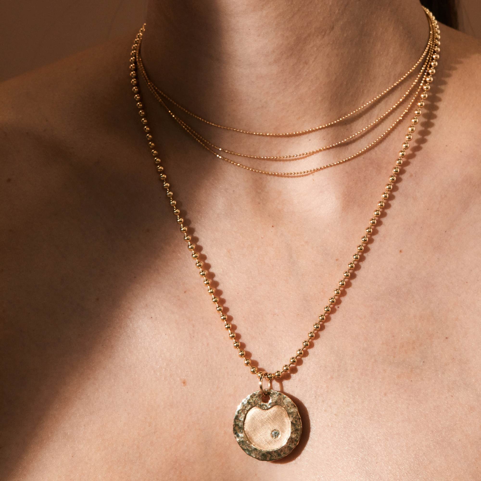 14k yellow gold three strand diamond cut ball chain stacked with the salk round pendant with hammered shiny edge and satin finish in the center with offset single white diamond on ball chain in lifestyle shot on model