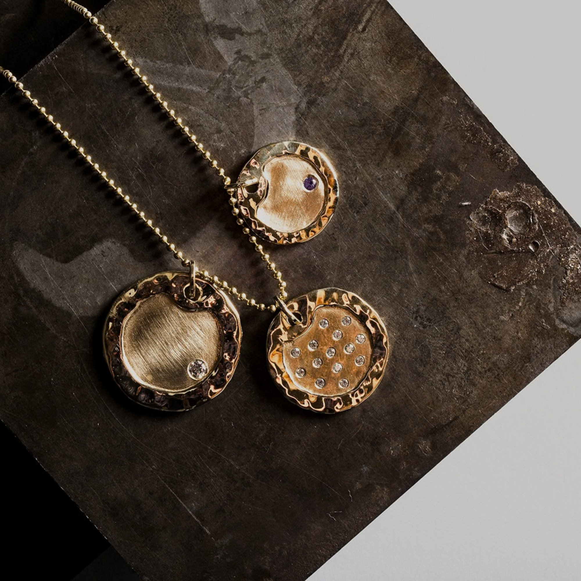 14k yellow gold round dogtag pendants with hammered shiny edge and brushed satin center with offset white diamond and offset colored gemstone next to the doff pendant which has multiple scattered white diamonds in lifestyle shot