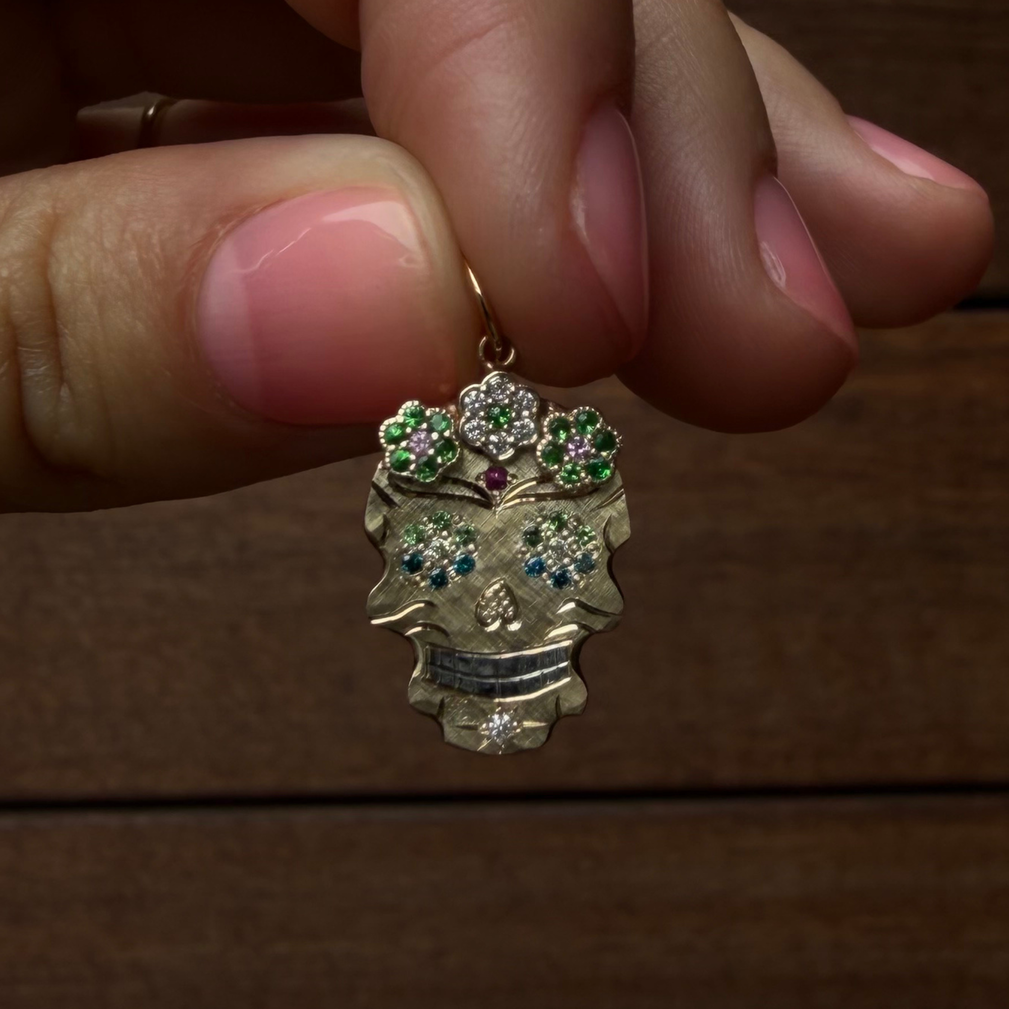 A close-up of a hand holding a gold sugar skull pendant decorated with green, blue, and white sapphires, set against a dark wooden background.