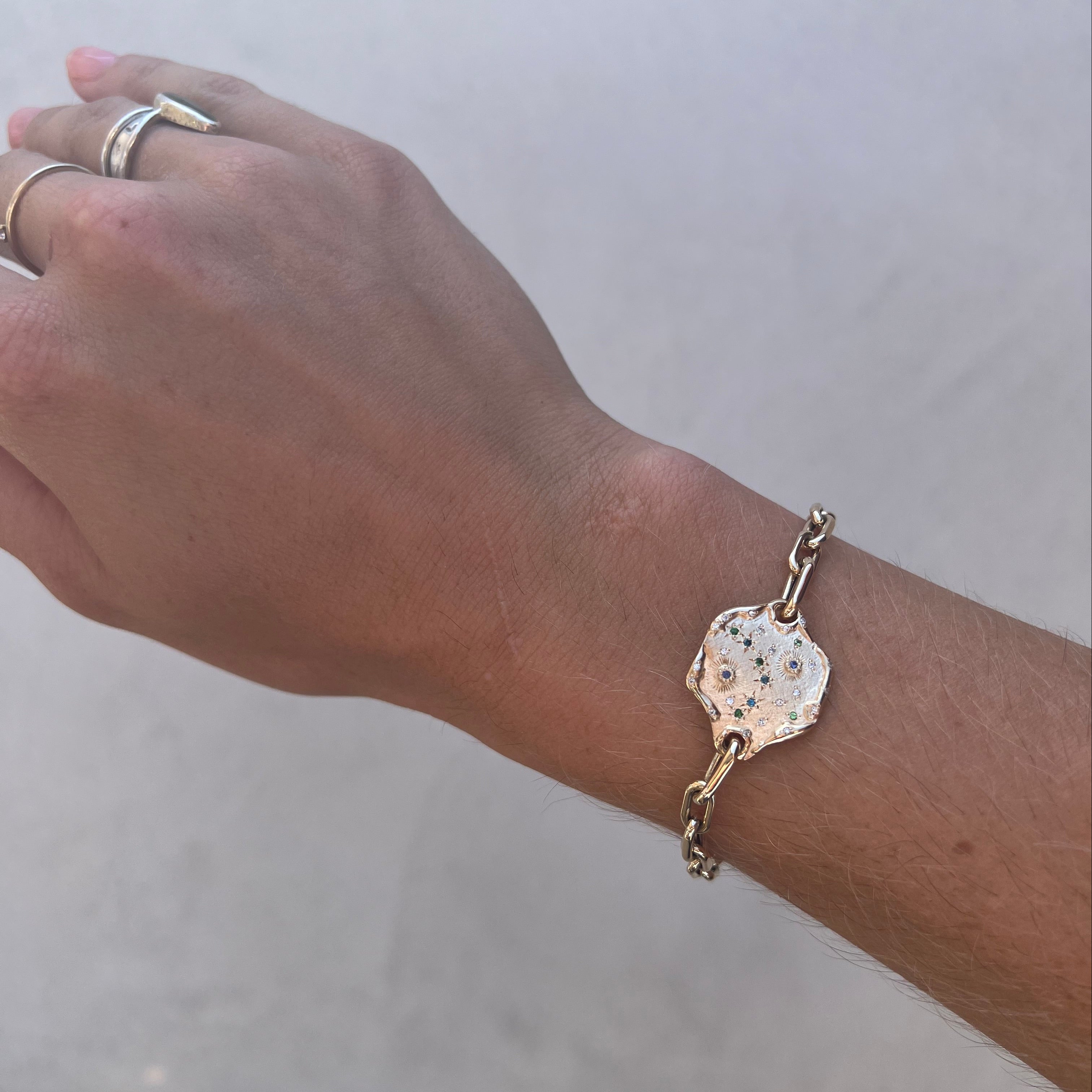 A gold-toned chain bracelet with a textured, organic-shaped charm featuring tiny black speckles, shown worn on a person's wrist against a neutral light gray background.