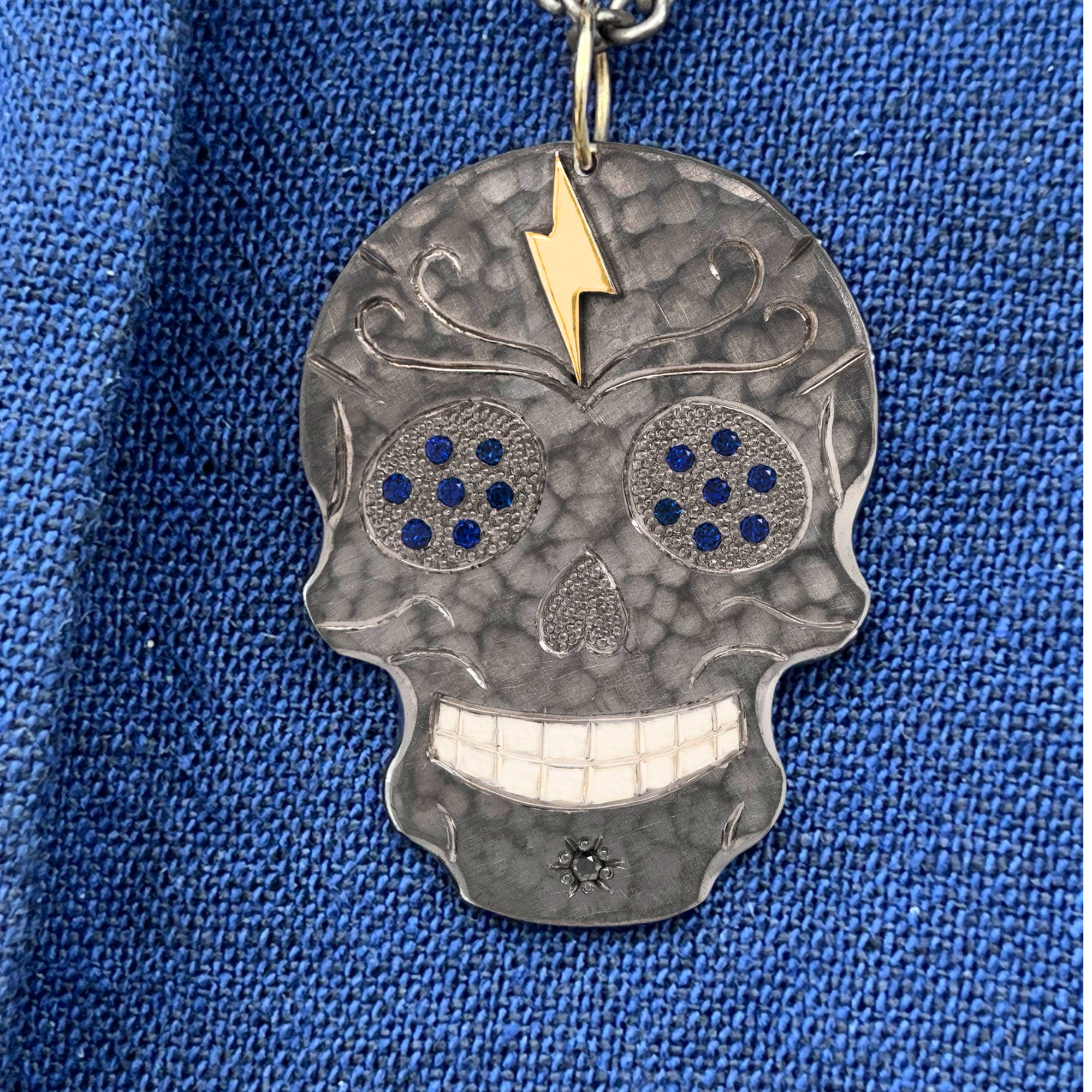 Skull-shaped pendant with blue gemstones and gold lightning bolt on a blue fabric background