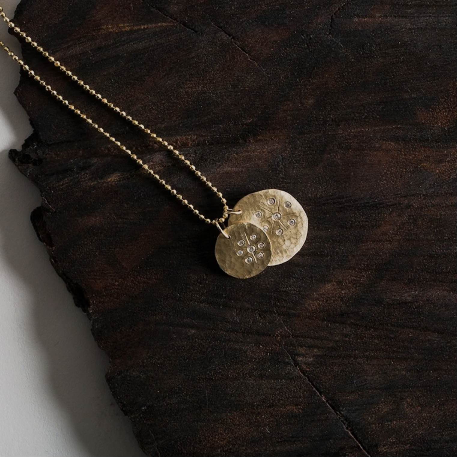 14k yellow gold round hammered pendant with white diamond starburst paired with larger round hammered pendant with white diamonds and hand etched starburst on ball chain on natural wood background