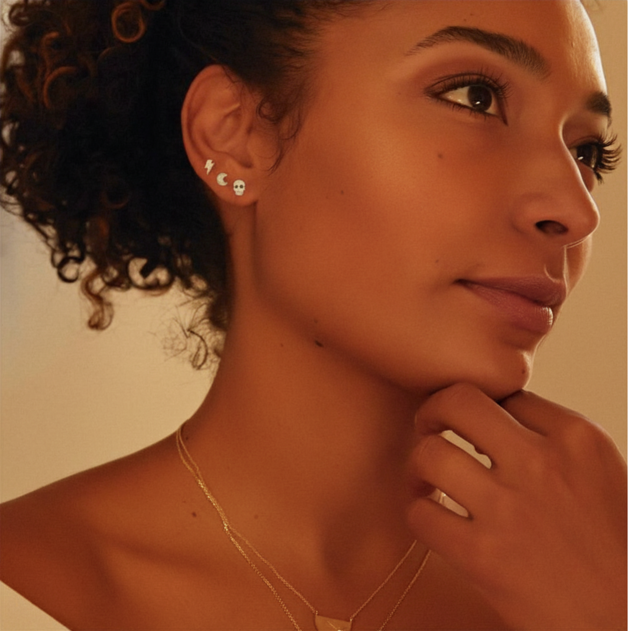 Close-up of a woman wearing tiny cutout shape stud earrings and a necklace with a warm, soft background.