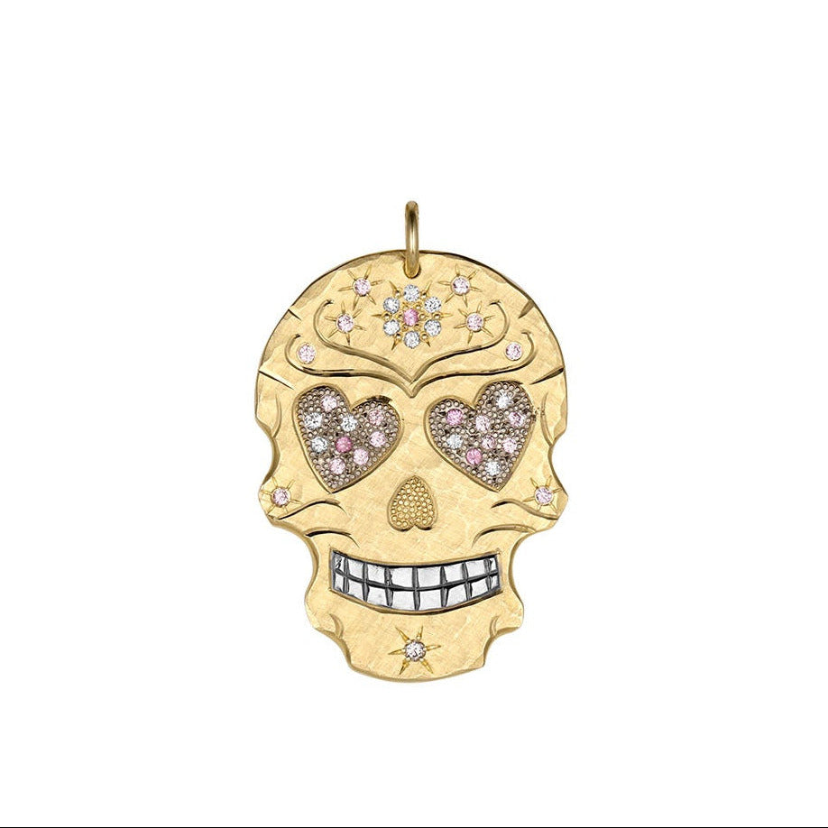 Large sized 14k yellow gold hand cut skull pendant featuring engraved details, shiny rhodium plated teeth with bead set gemstone floral and star motifs. Large heart shaped eyes are pave set with white diamonds and multi tone pink sapphires shown pendant only on white background.