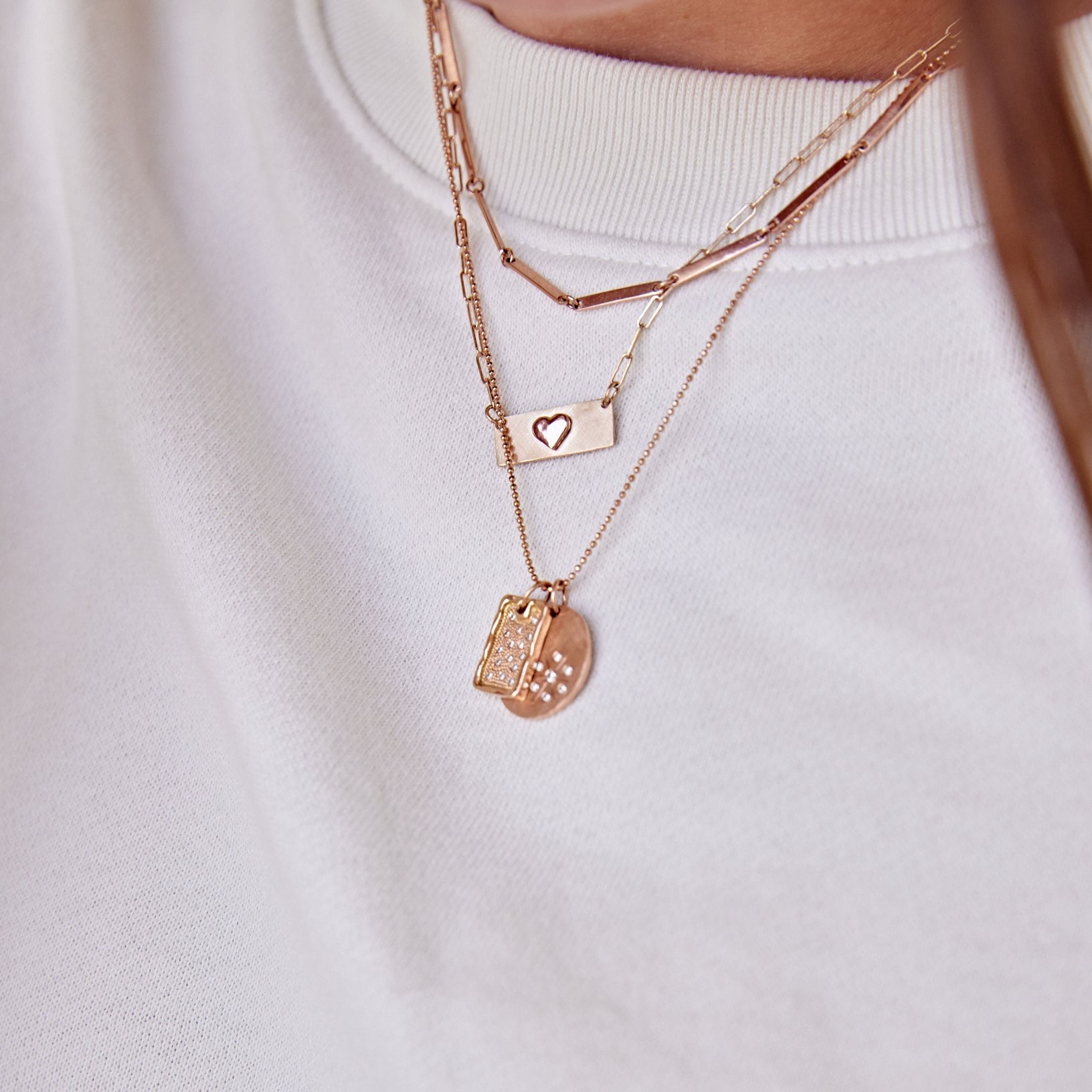 model in white sweatshirt wearing layered necklaces. Choker of flat bar links, dainty paperclip chain with raised heart tag necklace and a diamond ball chain with diamond dog tag and diamond disk medallion pendants.