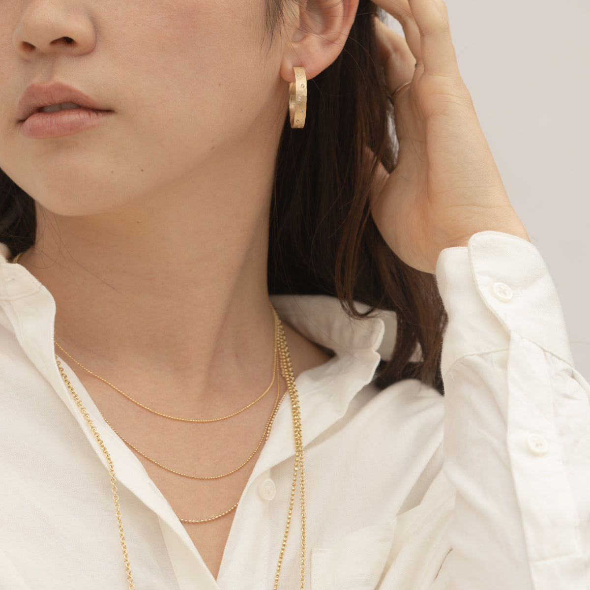 Model in white shirt wearing small sized wide hoops with scattered white diamonds and layered gold chain necklaces with white background