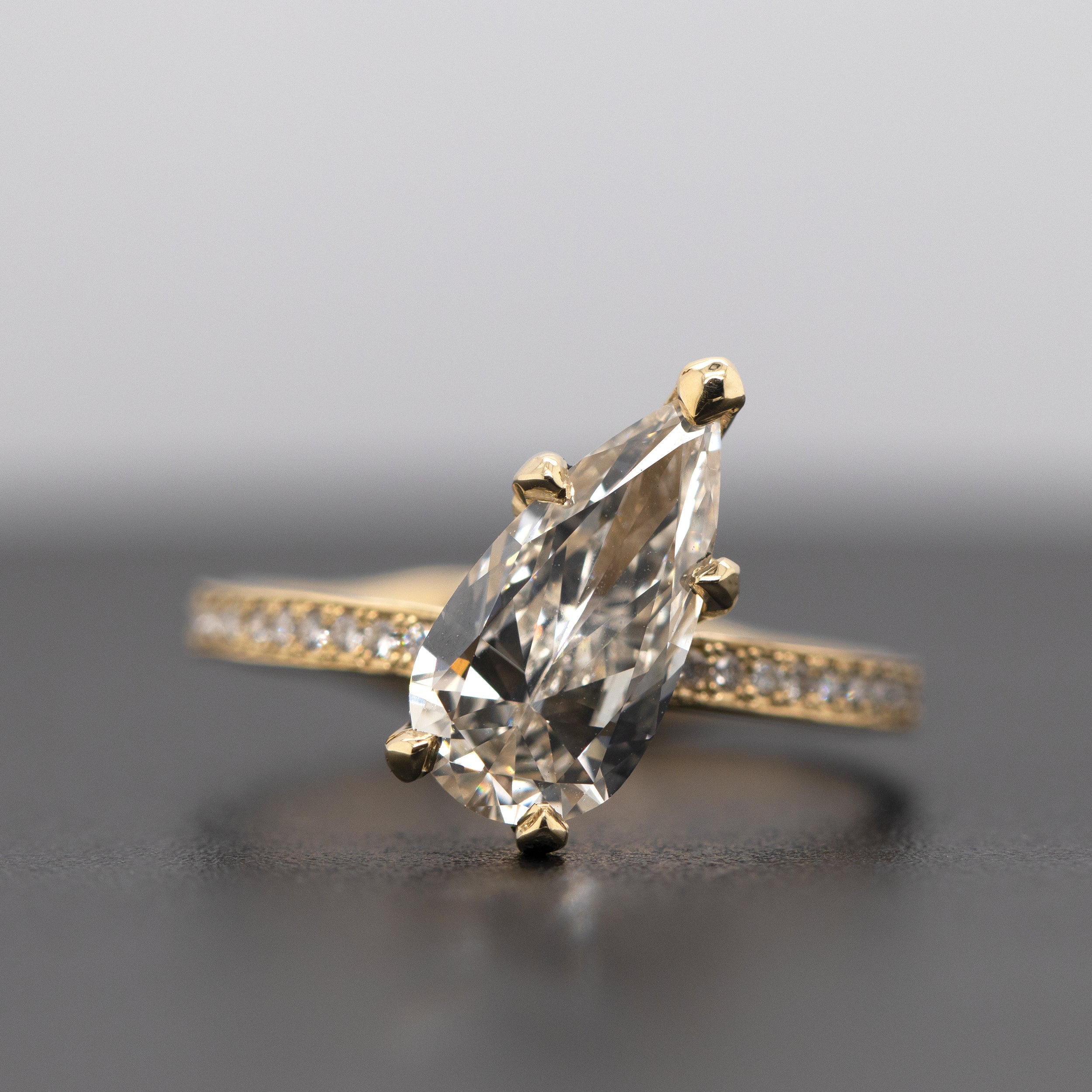 14k yellow gold ring featuring a tilted pear-shaped white diamond with five prongs and a half eternity band with pave set white diamonds and a thick comfort fit band on blurry surface background