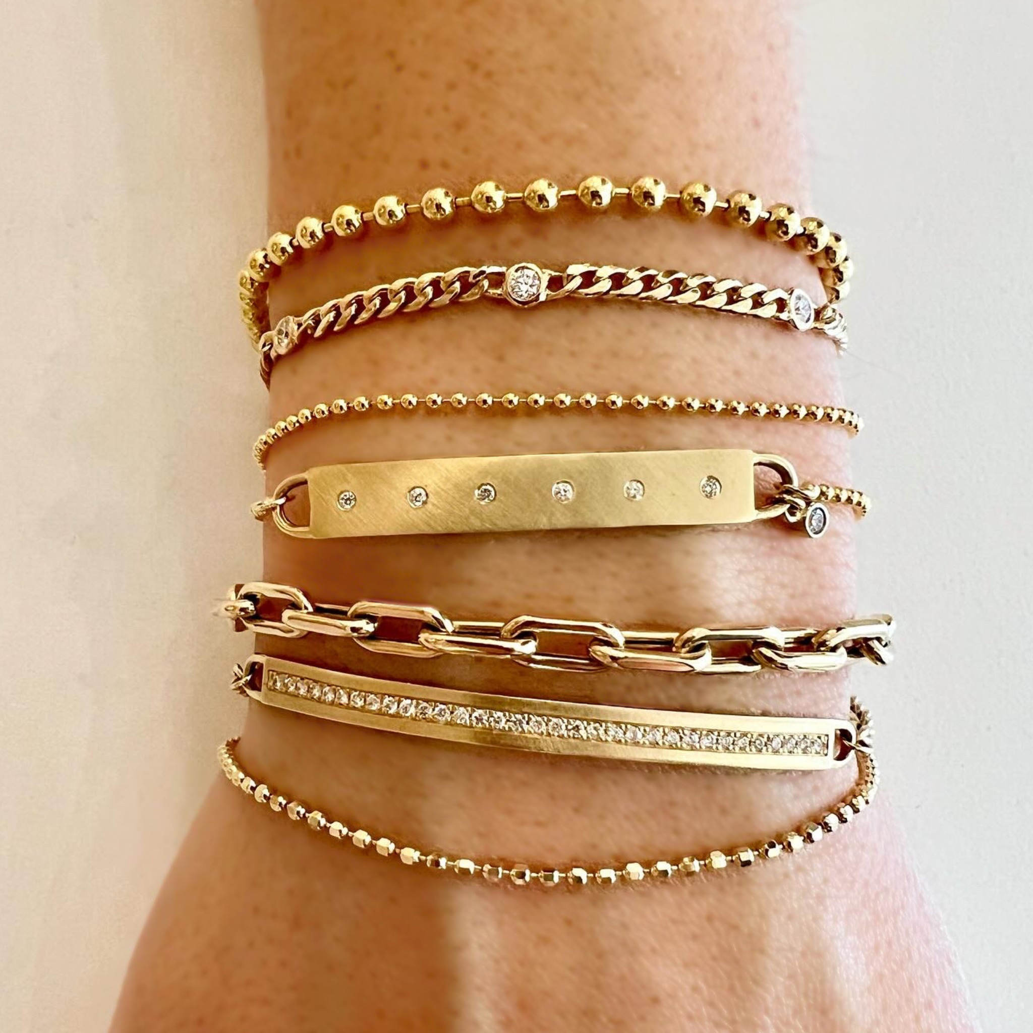 Close-up of a wrist wearing stacked gold bracelets with white diamond accents variety of types and textures on a neutral background