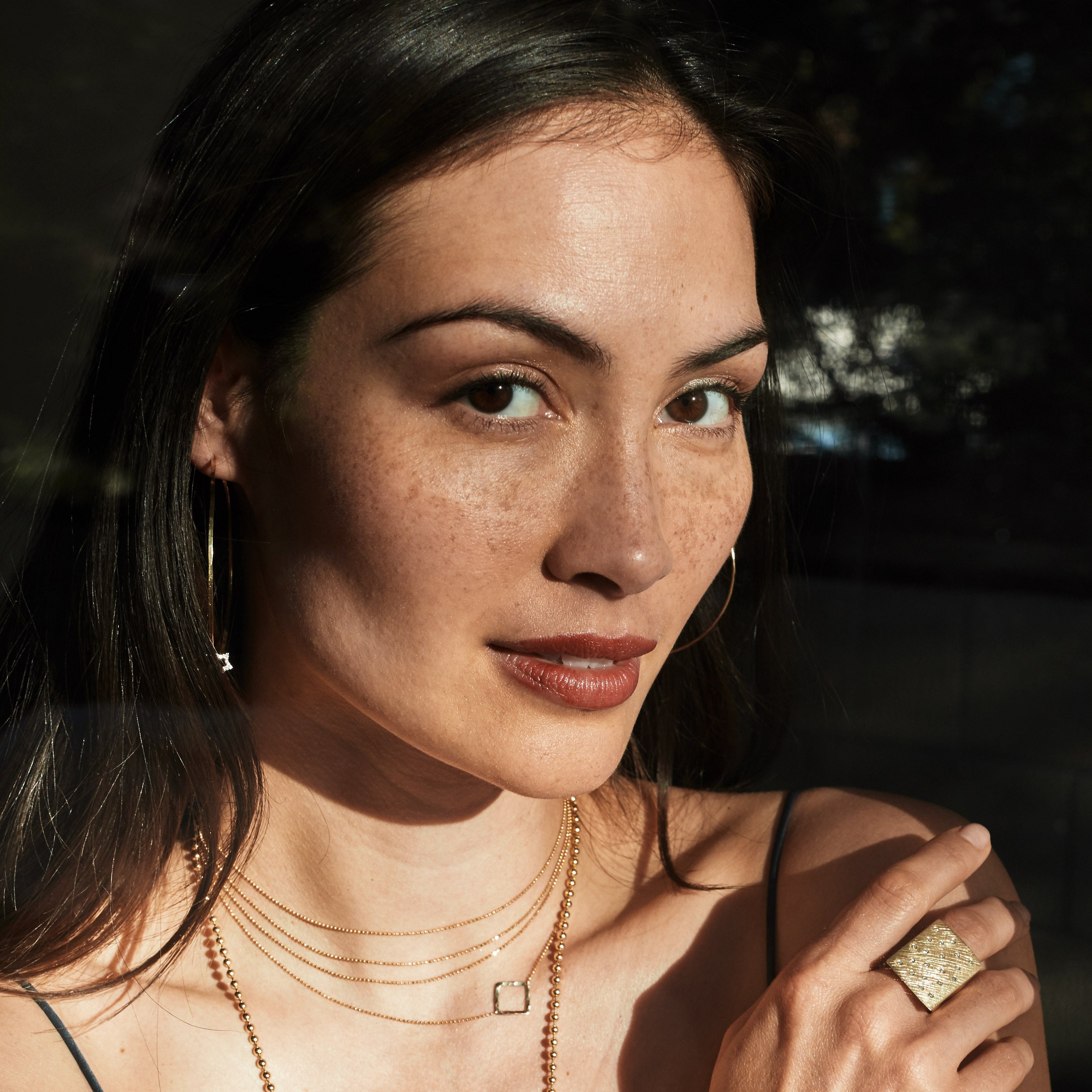 Woman wearing gold jewelry including NUMBER 1- 14k Yellow Gold Scattered White Diamond Signet Ring with Textured Finish, layered necklaces, hoop earrings  lifestyle shot dark background