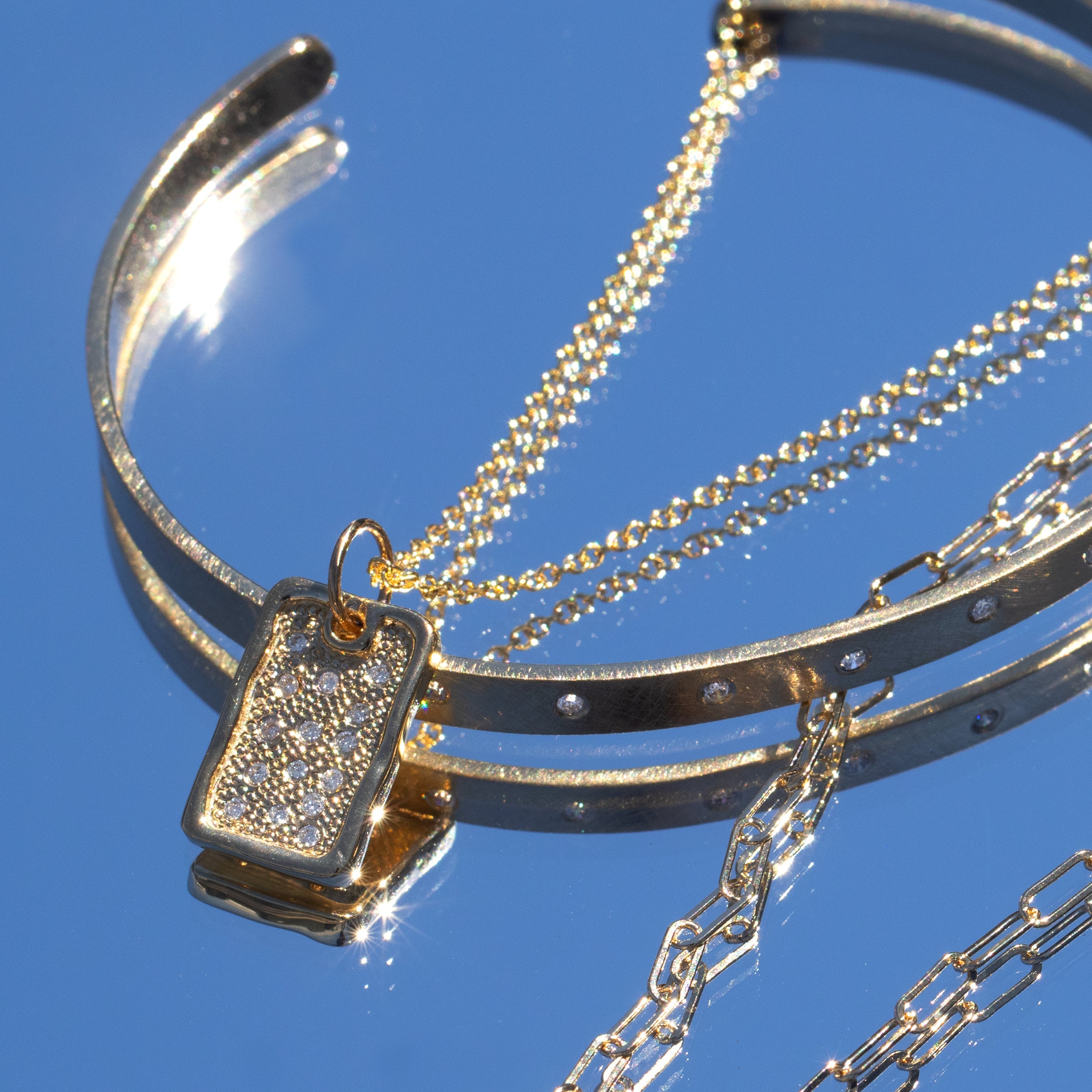 Group shot of 14 yellow gold jewelry on mirror with skye color background bangle and necklace featuring diamond studded dog tag
